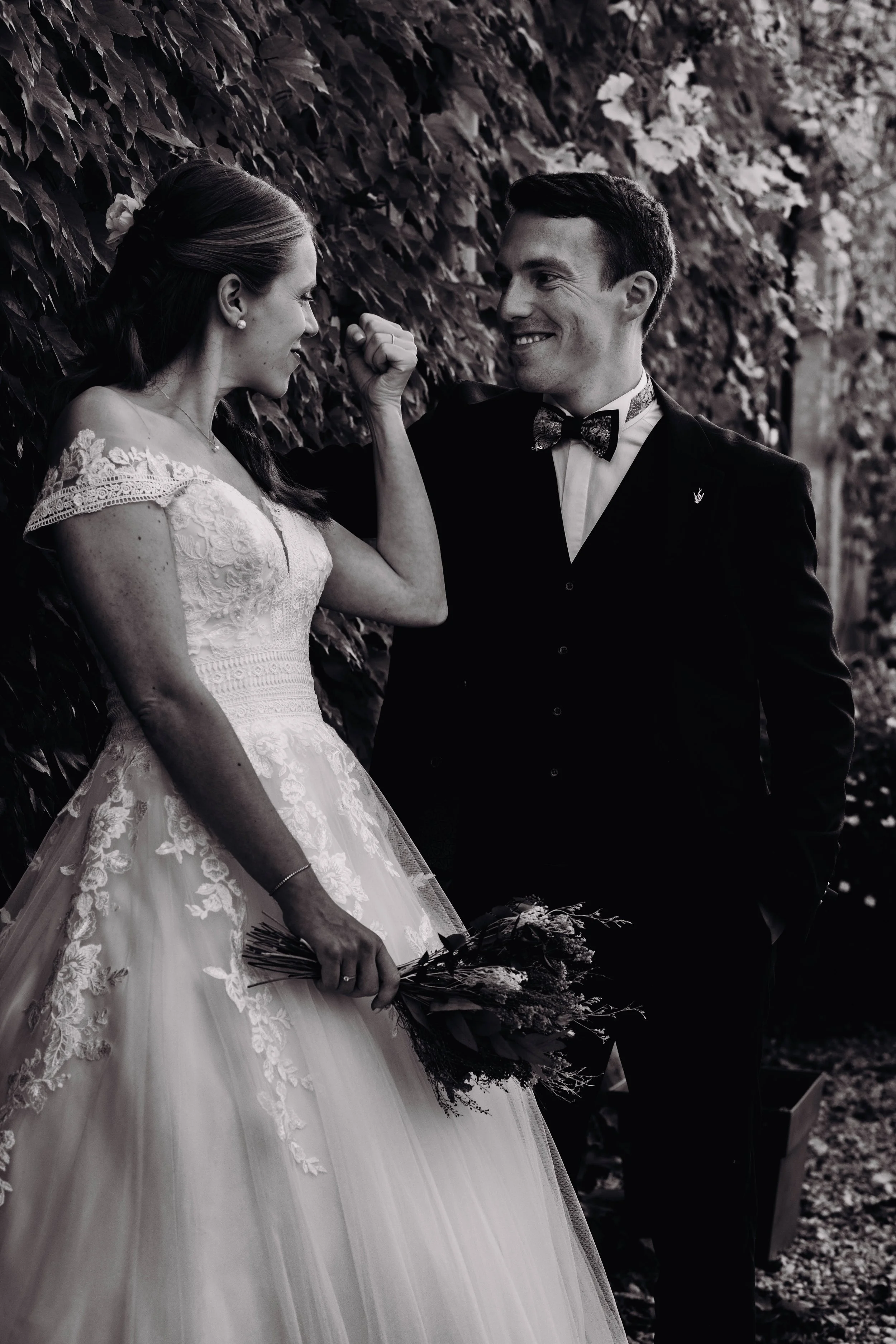 Photo en noir et blanc d'un couple de mariés lors d'un mariage, la mariée portant une robe en dentelle et tenant un bouquet, ils échangent un regard complice avec la mariée au manoir du Thouron proche de Toulouse. 