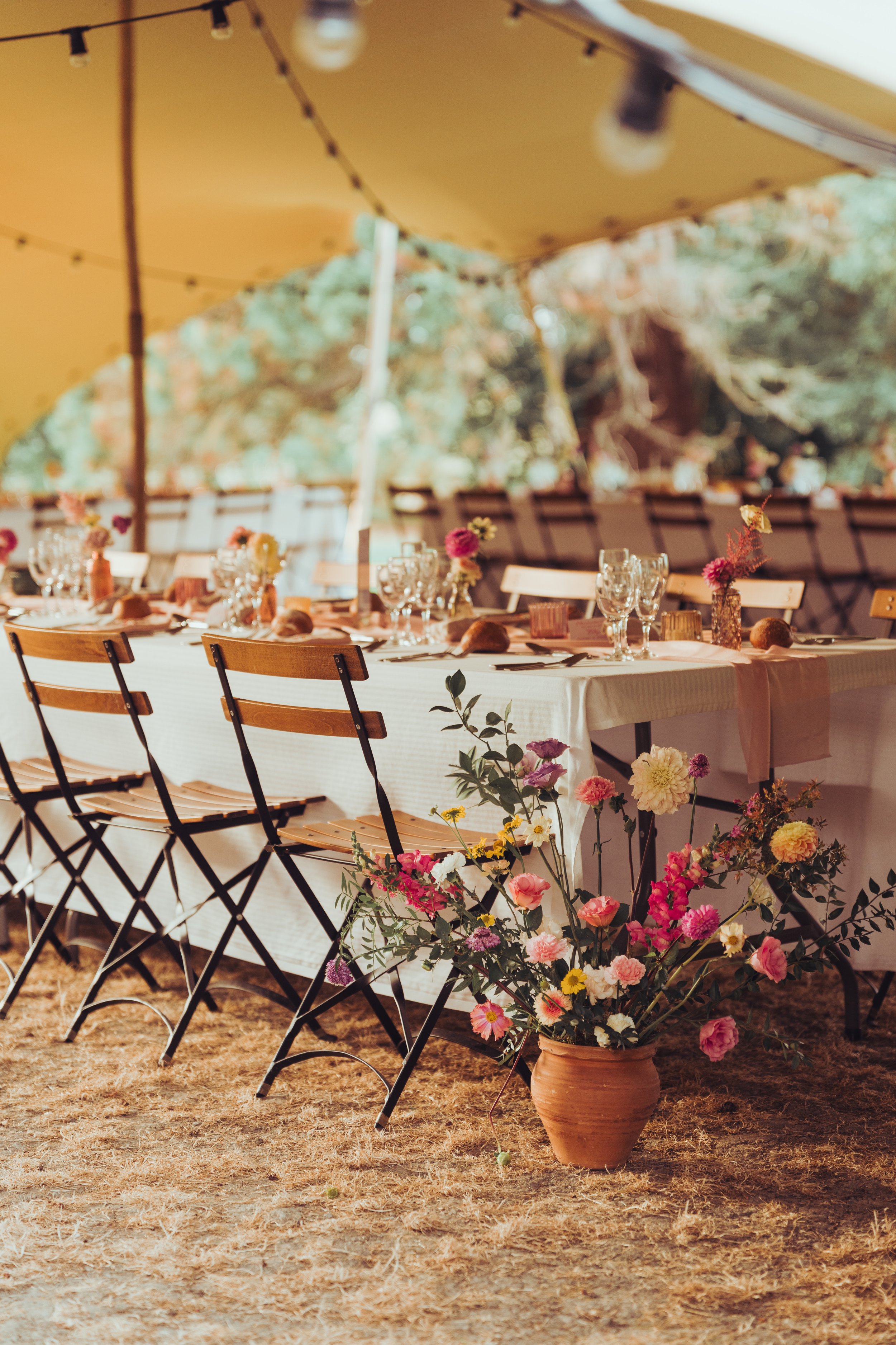 Une table dressée pour un mariage en plein air au chateau de Fajac la Relenque, décorée avec des fleurs et des verres par Cespetitsriendéco, sous une grande tente. En premier plan, un pot de fleurs avec des roses et autres fleurs colorées.
