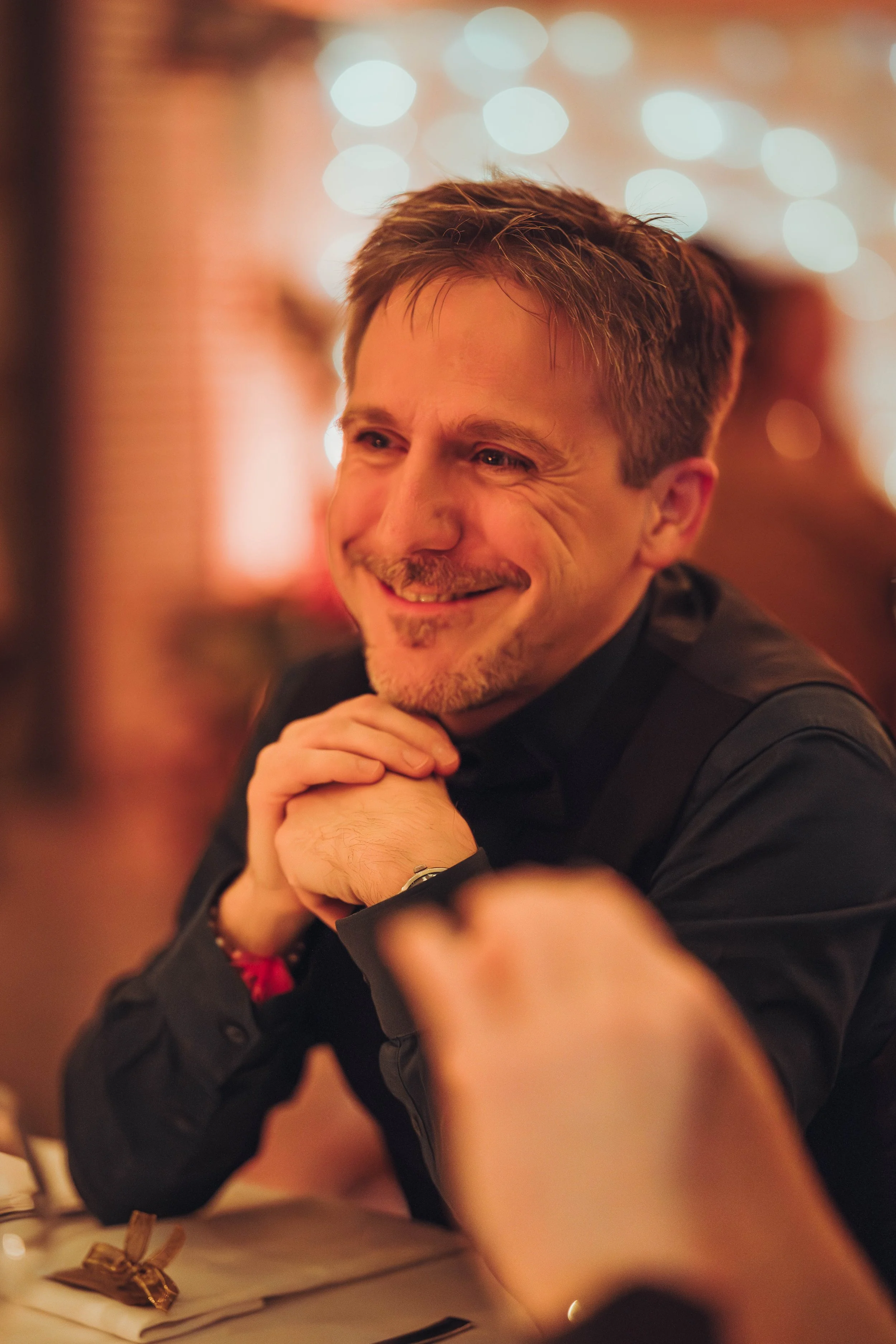 Un homme souriant avec une barbe courte, vêtu d'une chemise noire, assis à une table dans un endroit éclairé par des lumières chaudes dans un décor chaleureux au domaine de Gailhaguet proche de Toulouse.