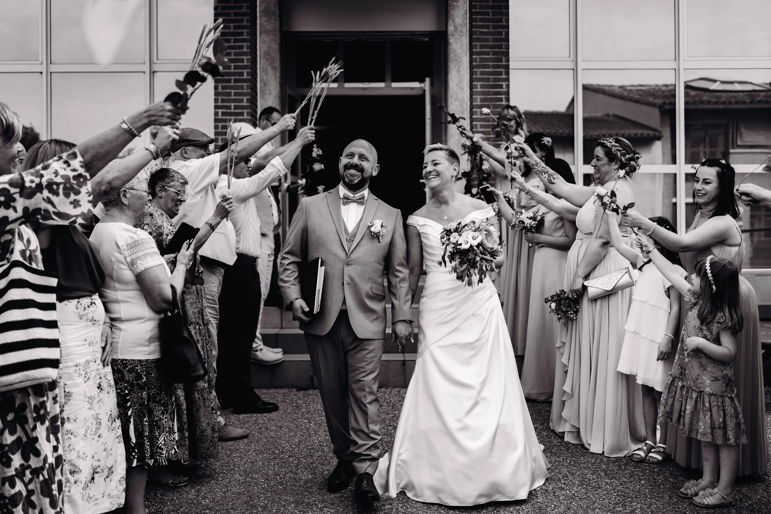Photo en noir et blanc d'un couple de mariés marchant main dans la main à la sortie de la mairie de Fonsorbes, proche de Toulouse, entourés d'amis et de famille leur lançant des fleurs pour célébrer leur mariage.