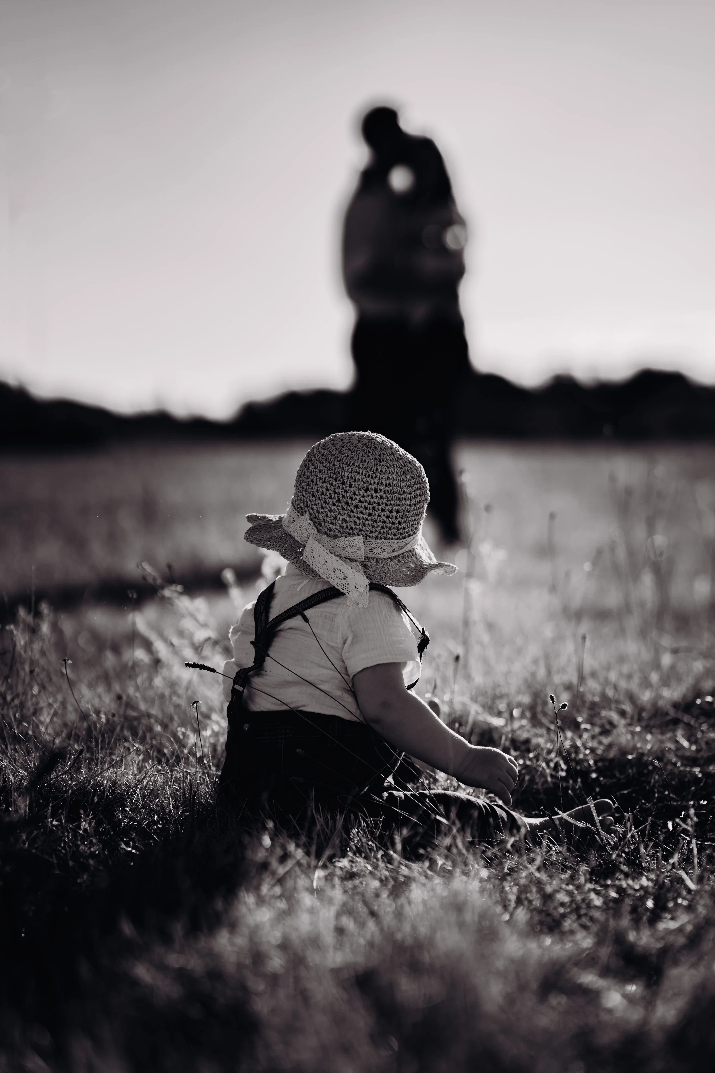 Une jeune enfant assise dans l'herbe portant un chapeau en crochet, regardant un homme distant qui est flou en arrière-plan, en noir et blanc.
