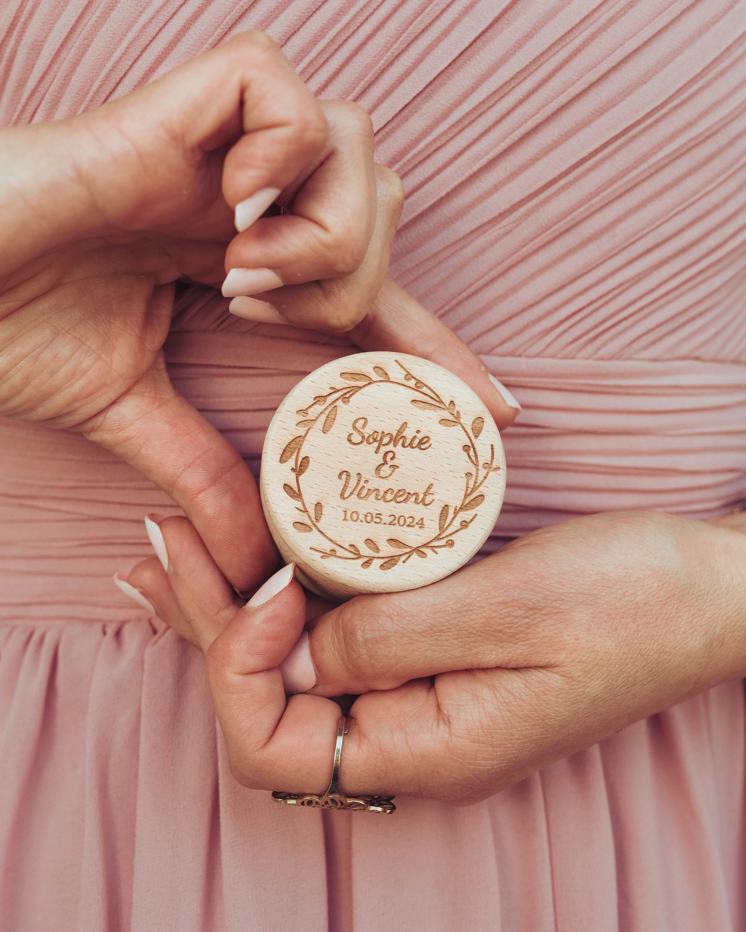 Mains tenant un petit boîtier en bois gravé avec des noms, une date et une décoration florale, devant une robe rose plissée. Souvenir pour les invités d'un mariage proche de Toulouse.