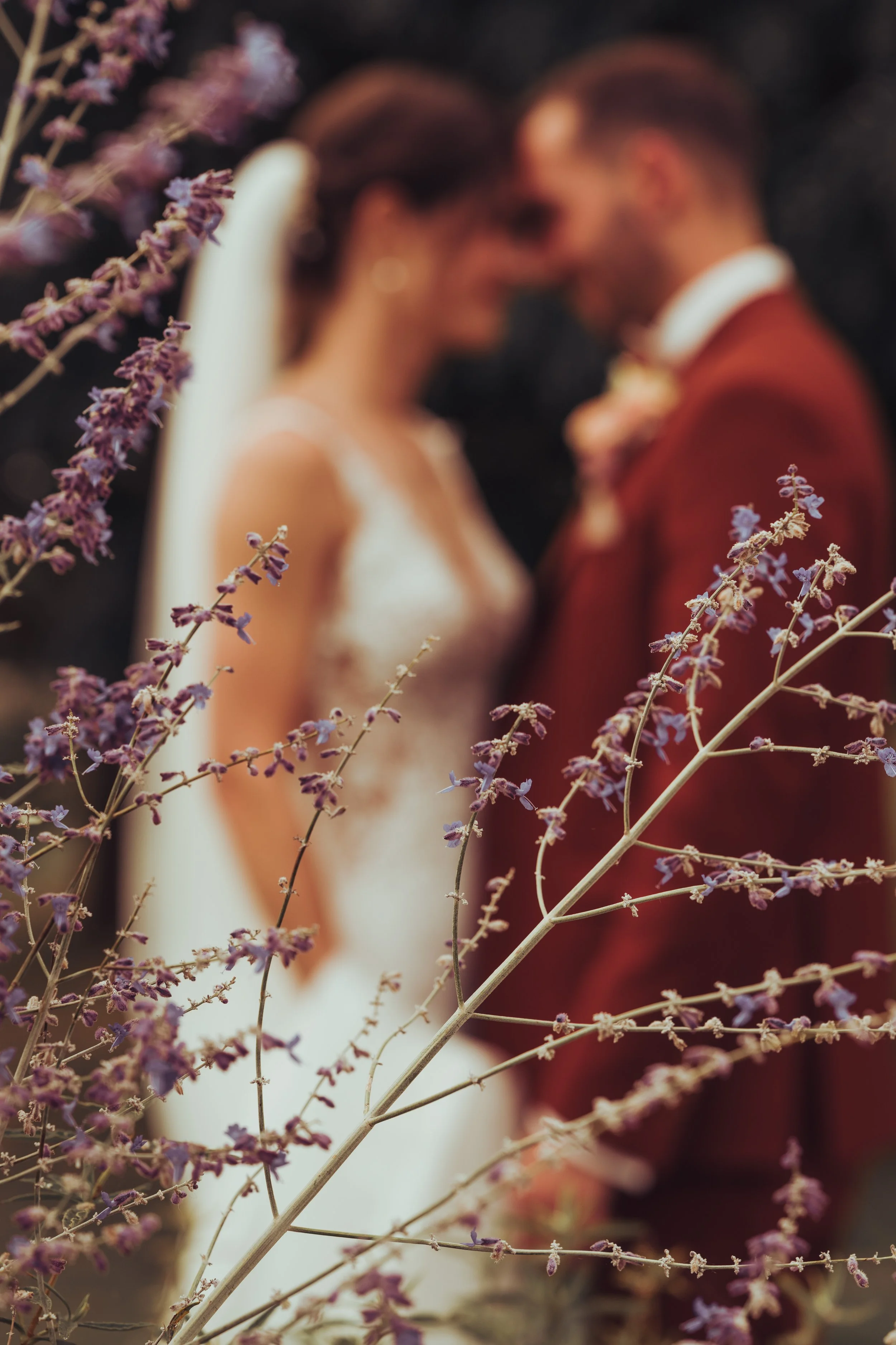 Une photo floue intentionnel d'un couple de mariés en veste rouge et robe de mariée blanche, se tenant très près, à l'extérieur, avec des fleurs violettes au premier plan au domaine de Fajac la relenque, proche de Toulouse