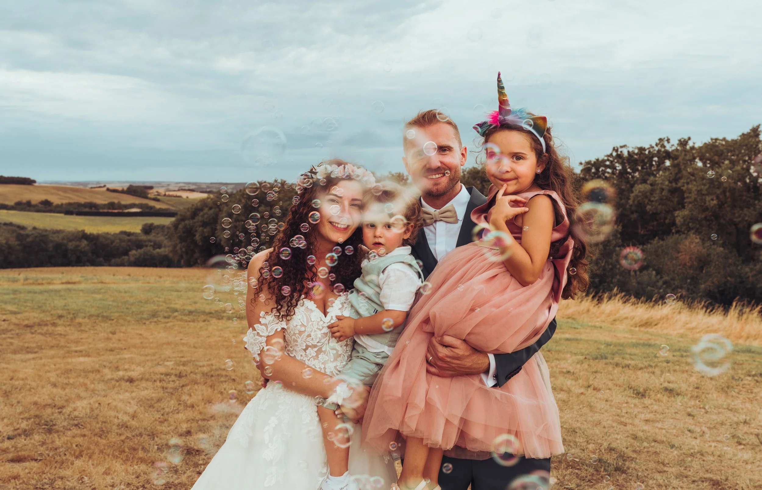 Séance photo de famille en plein air lors d'un mariage à la Métairie du Fusté, avec des bulles de savon flottant autour, dans un champ avec les collines du gers en arrière-plan.