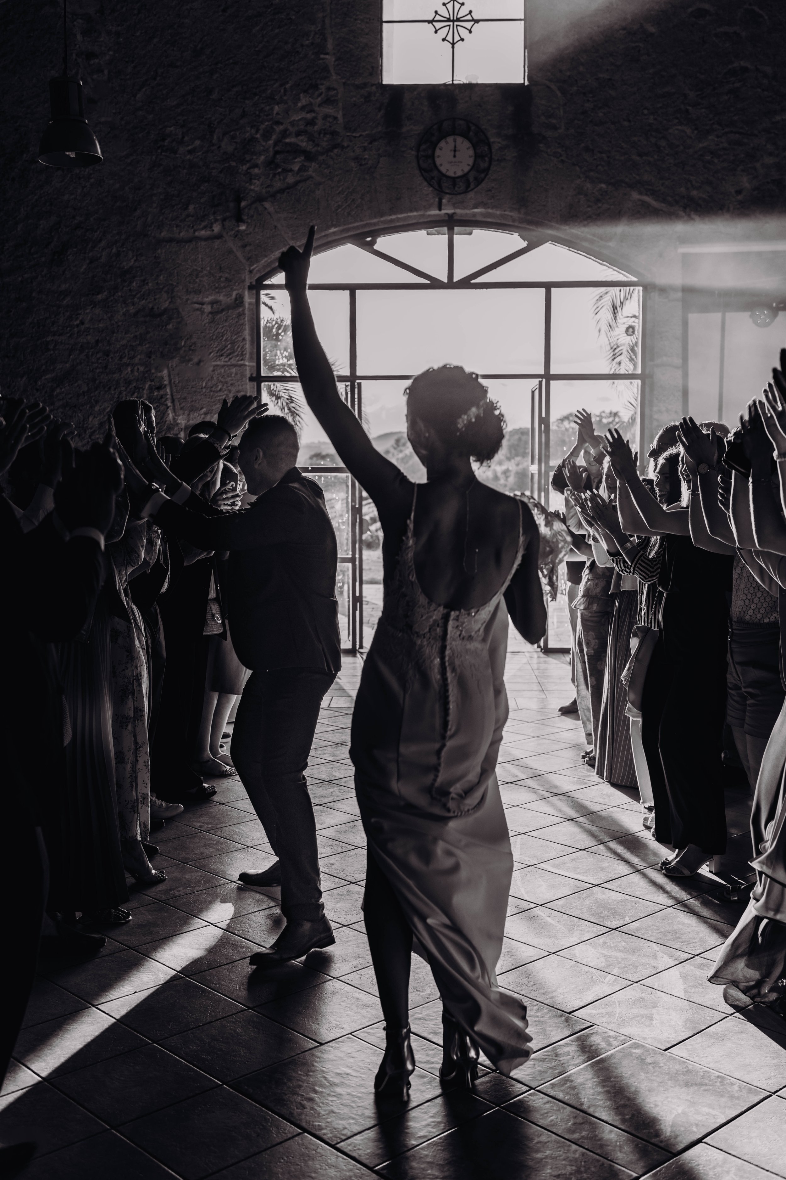 La mariée danse au centre d'une salle entourée de personnes applaudissant, en silhouette contre la lumière du soleil entrant par une grande fenêtre lors de son mariage proche de Béziers, Occitanie. 