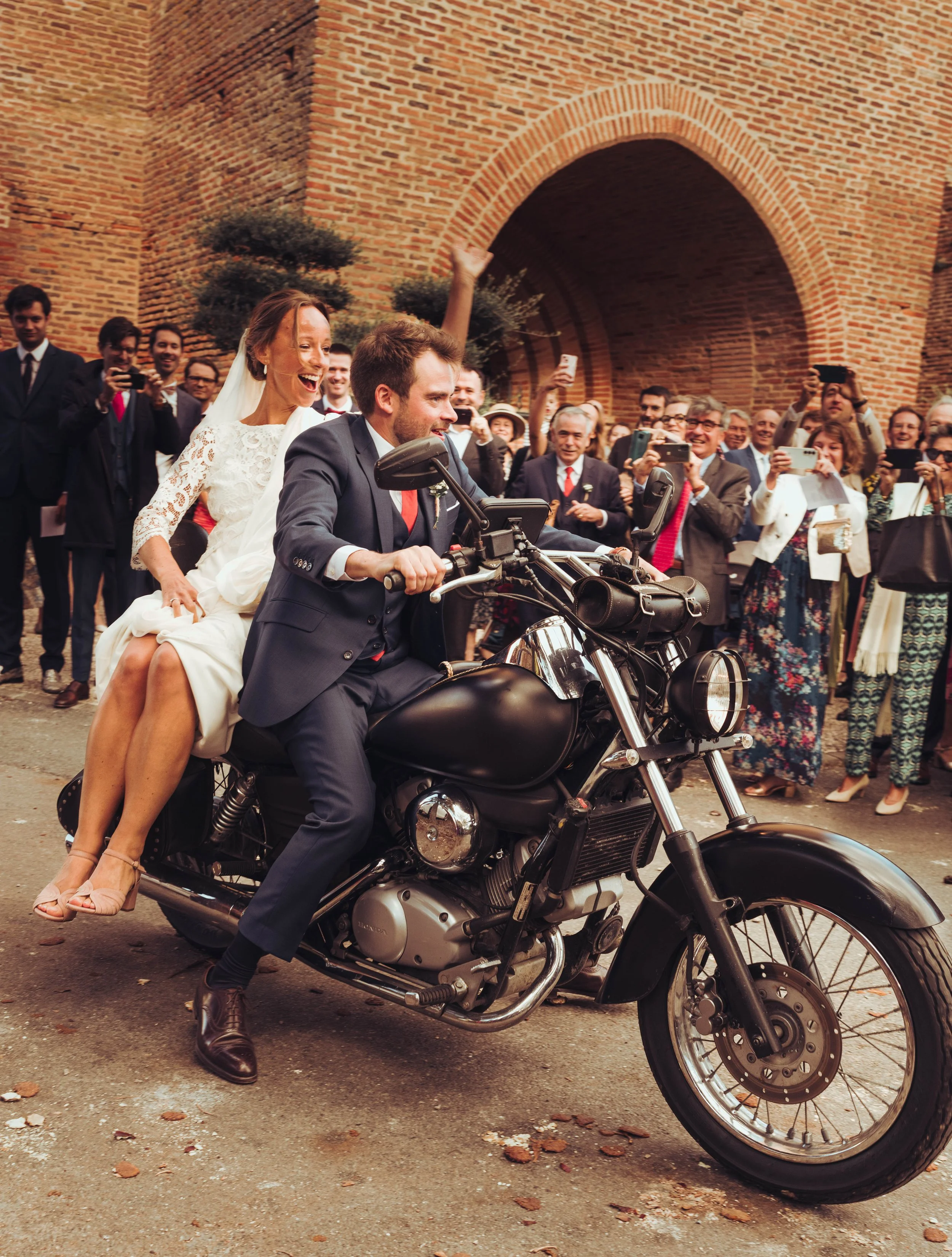 Un couple de mariés en costume et robe blanche sur une moto à la sortie de leur cérémonie religieuse proche du manoir de Lisle, entouré de nombreux invités qui prennent des photos, avec une architecture en briques en arrière-plan.