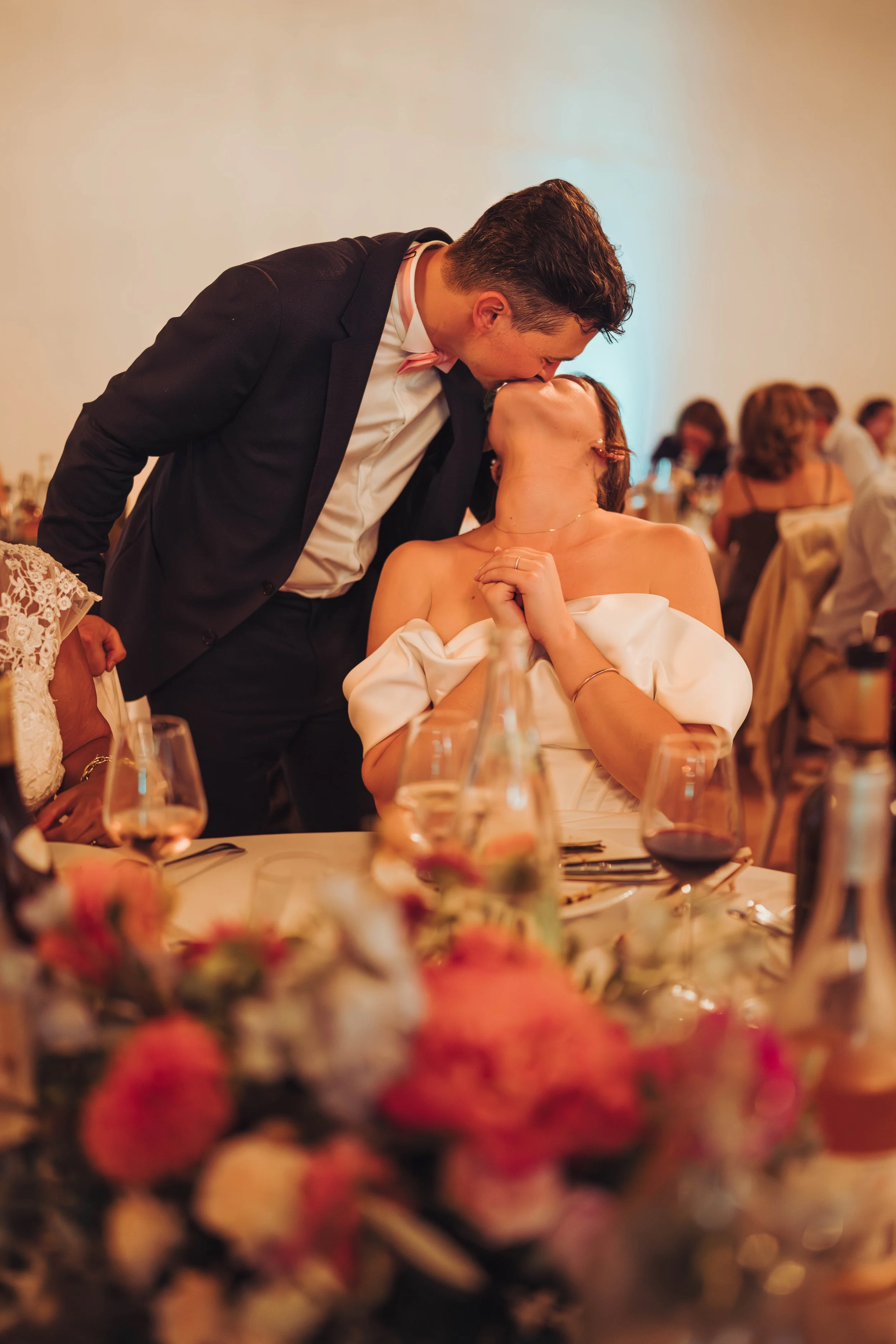 Un couple s'embrasse lors de leur réception de mariage au manoir de Lisle, près de Toulouse, en Occitanie, dans une salle décorée, avec d'autres invités en arrière-plan.