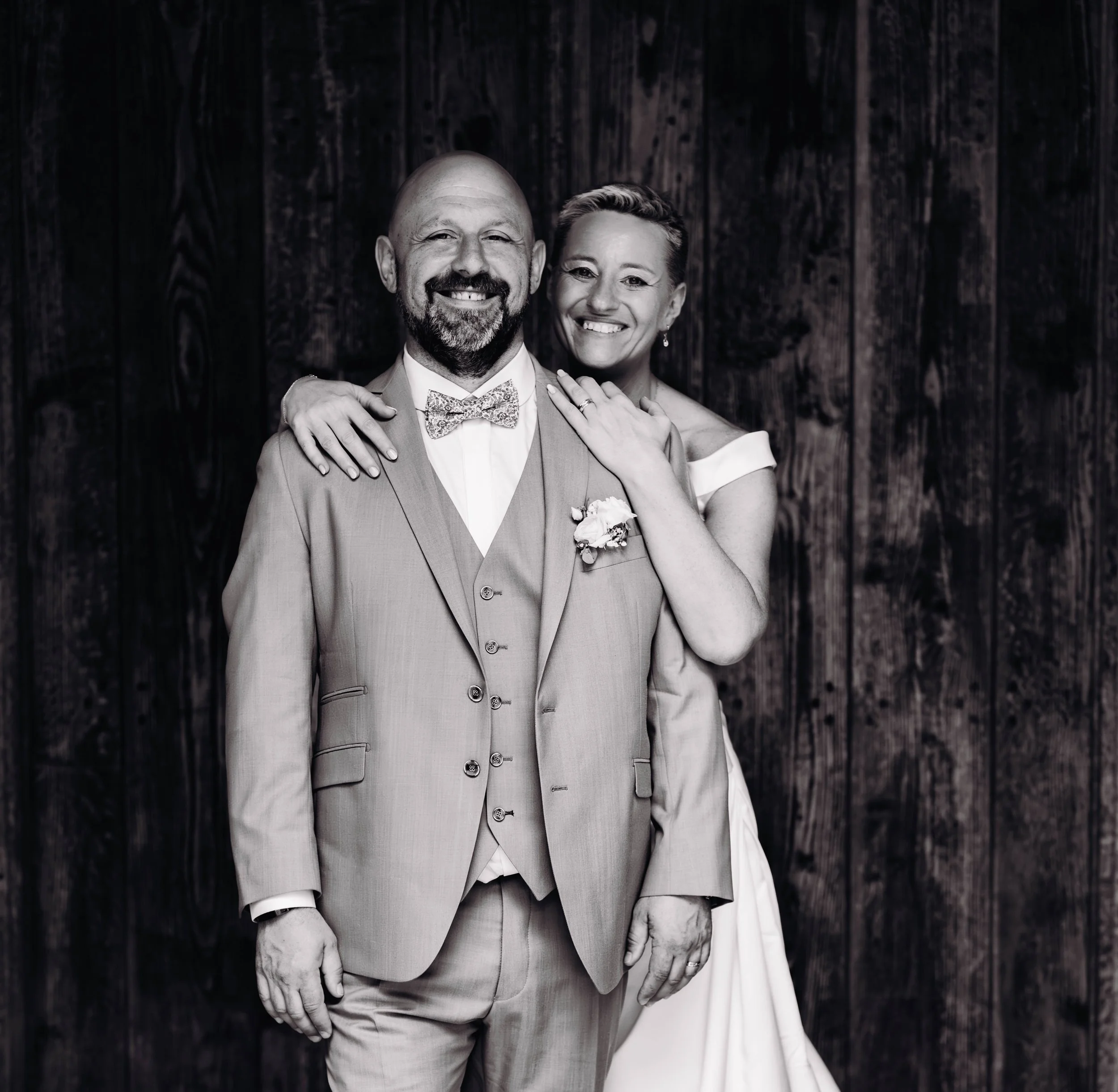Un couple lors de leur mariage, en costume pour l'homme et robe de mariée blanche pour la femme, souriant devant un fond en bois à l'entrée de l'église avant leur cérémonie religieuse proche de Toulouse.