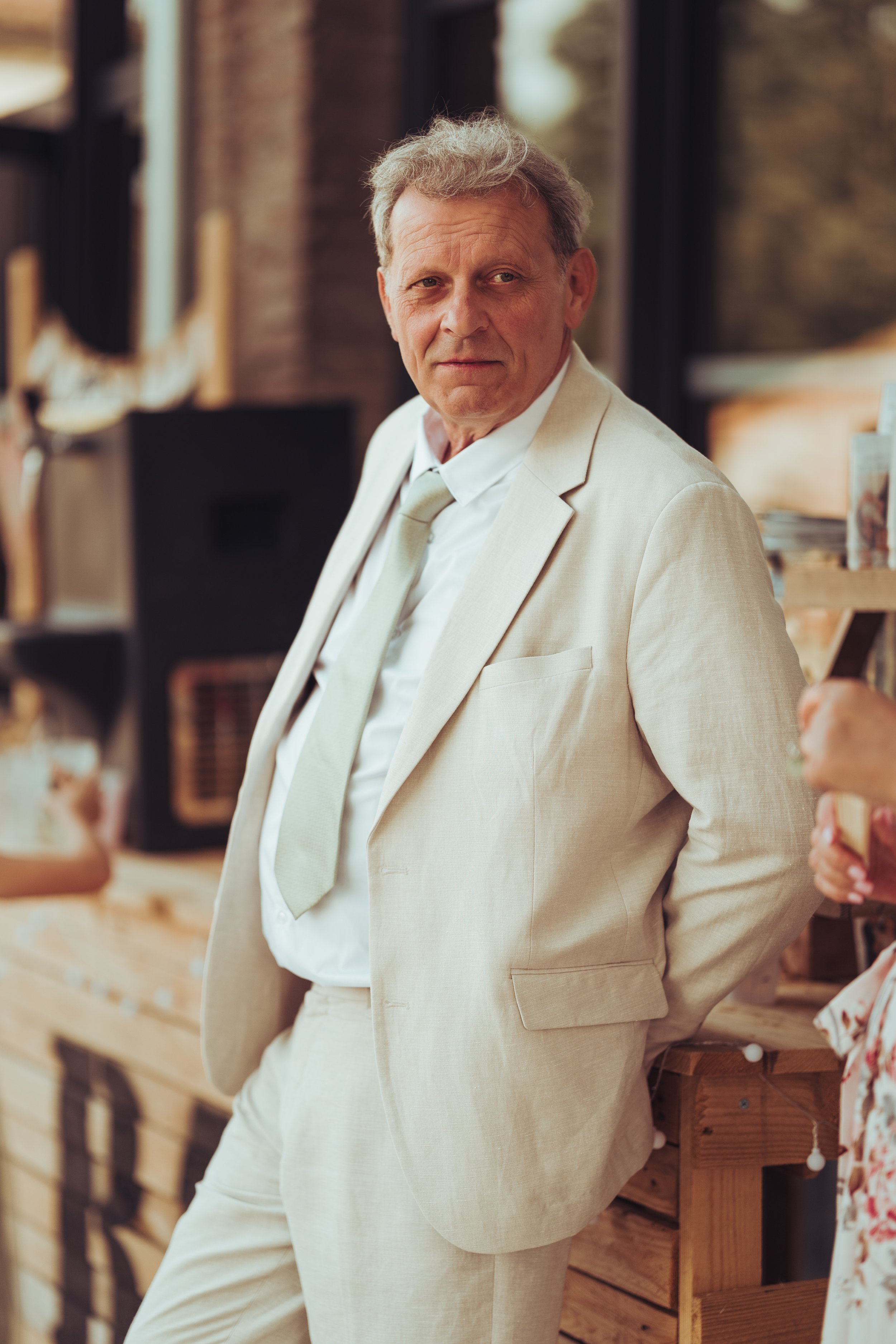 Père de la mariée à la métairie du Fusté proche de Toulouse, en Occitanie avec chemise blanche et cravate beige, debout avec les mains derrière le dos dans un lieu intérieur avec meubles en bois et grandes fenêtres
