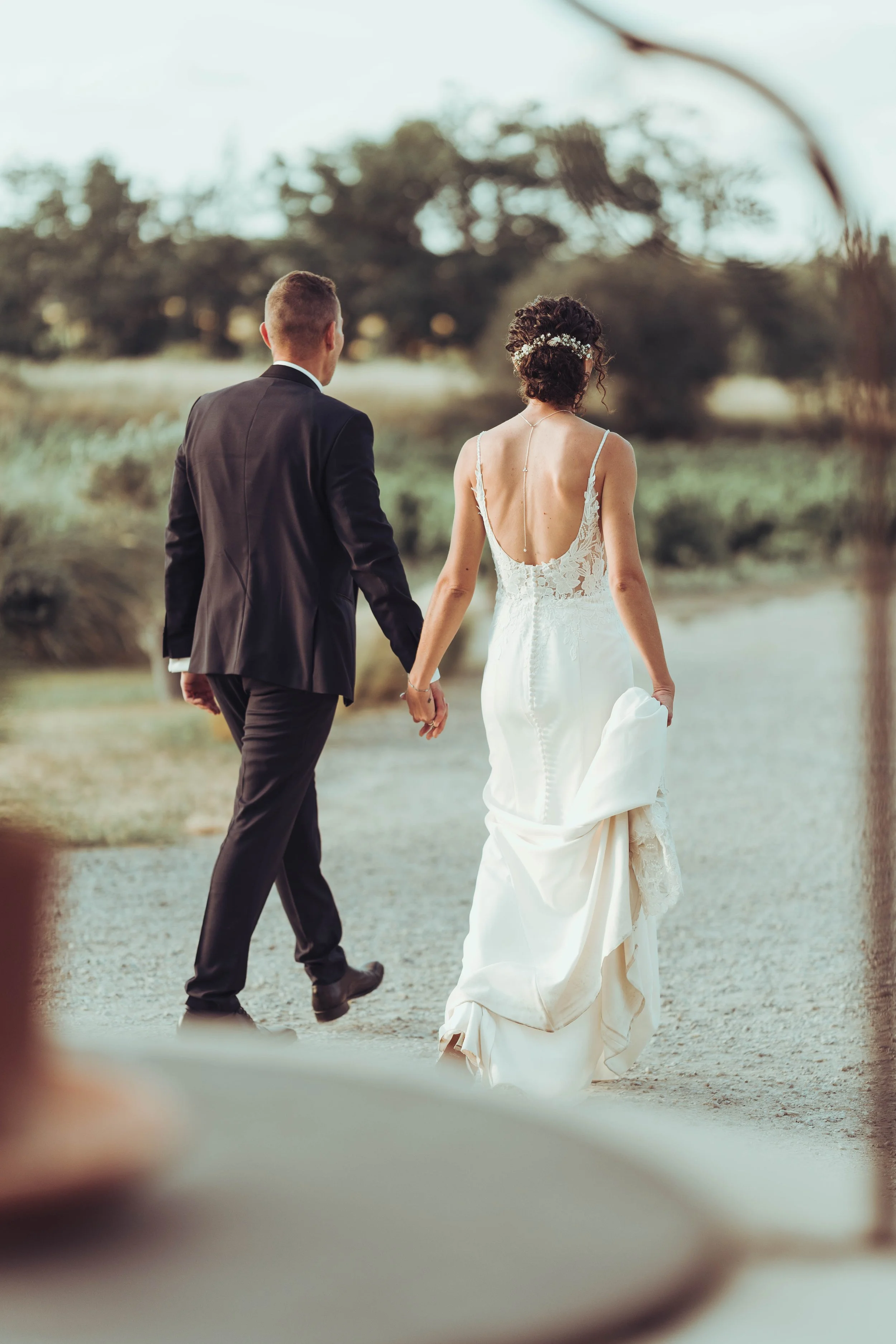 Un couple marié marche main dans la main dans un paysage extérieur naturel à Béziers, proche de Toulouse, la mariée porte une robe de mariée blanche et le marié un costume noir.