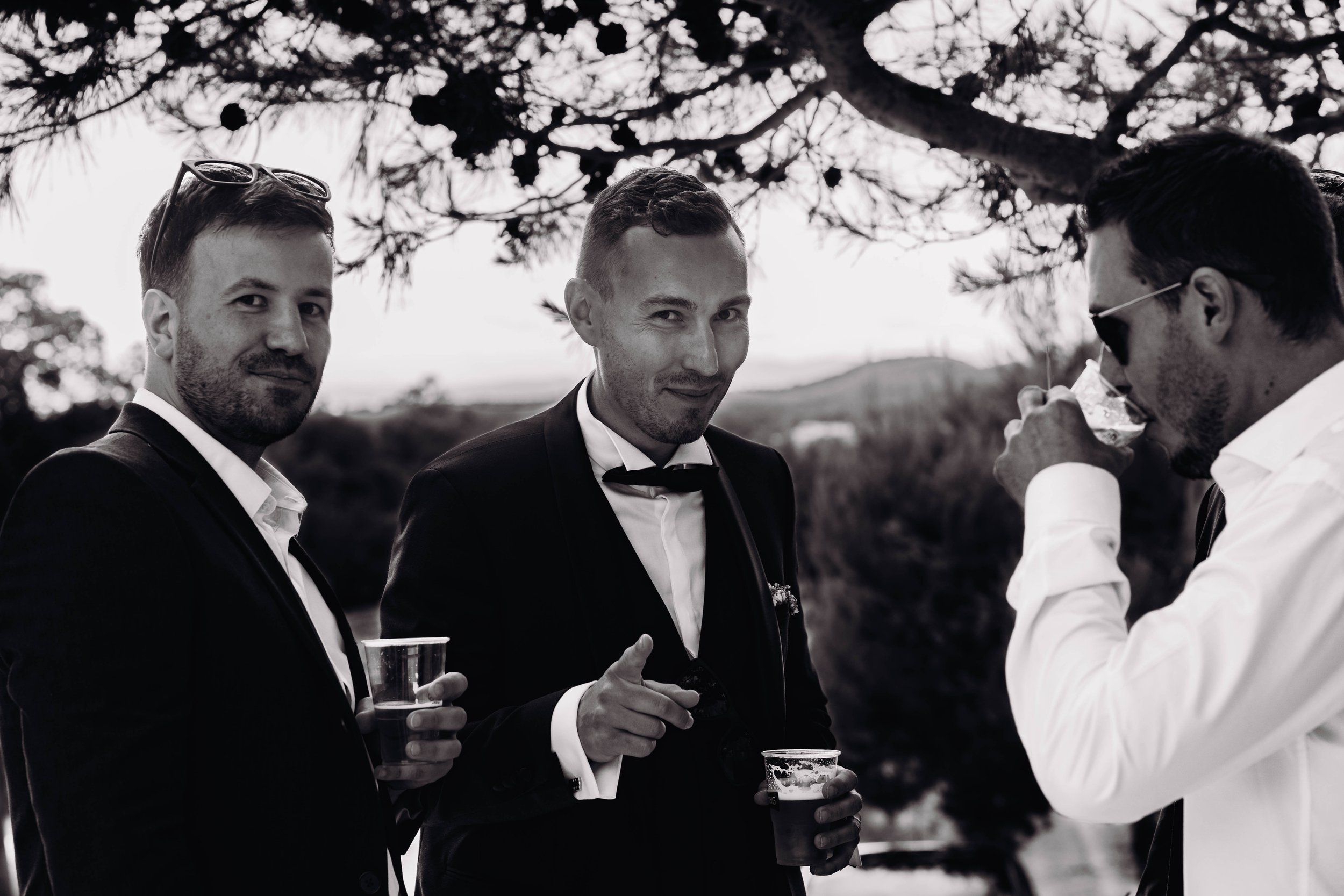 Trois hommes en costumes noirs et blancs lors d'un mariage à Béziers en Occitanie l'un d'eux tenant un verre, discutent lors d'une occasion en plein air sous un arbre.