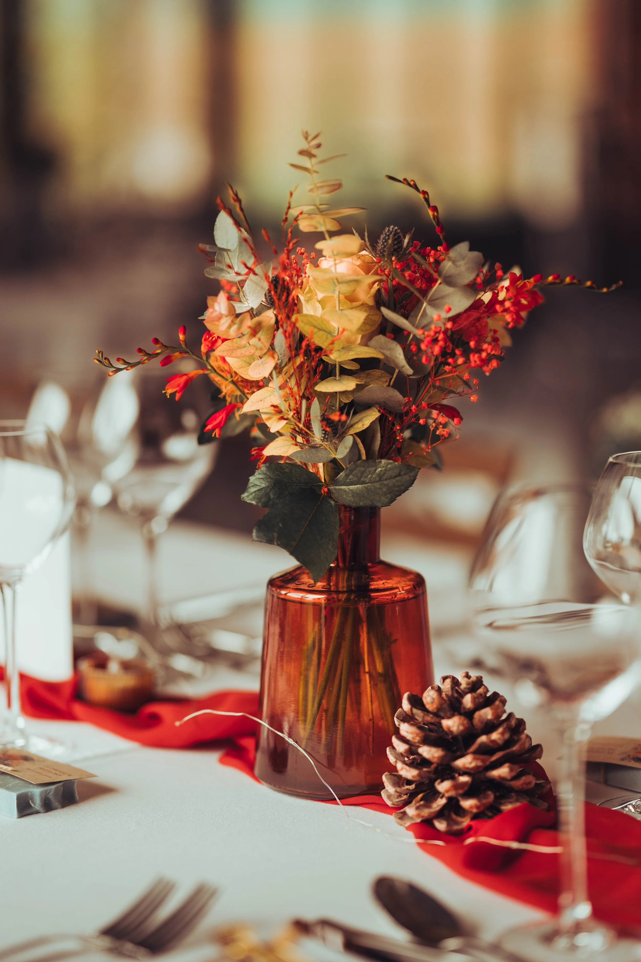 Centre de table de mariage avec un bouquet de fleurs sèches dans un vase en verre brun, décoré d'une branche de baies rouges, sur une table avec une mèche de gui et une pomme de pin, pour une décoration festive.