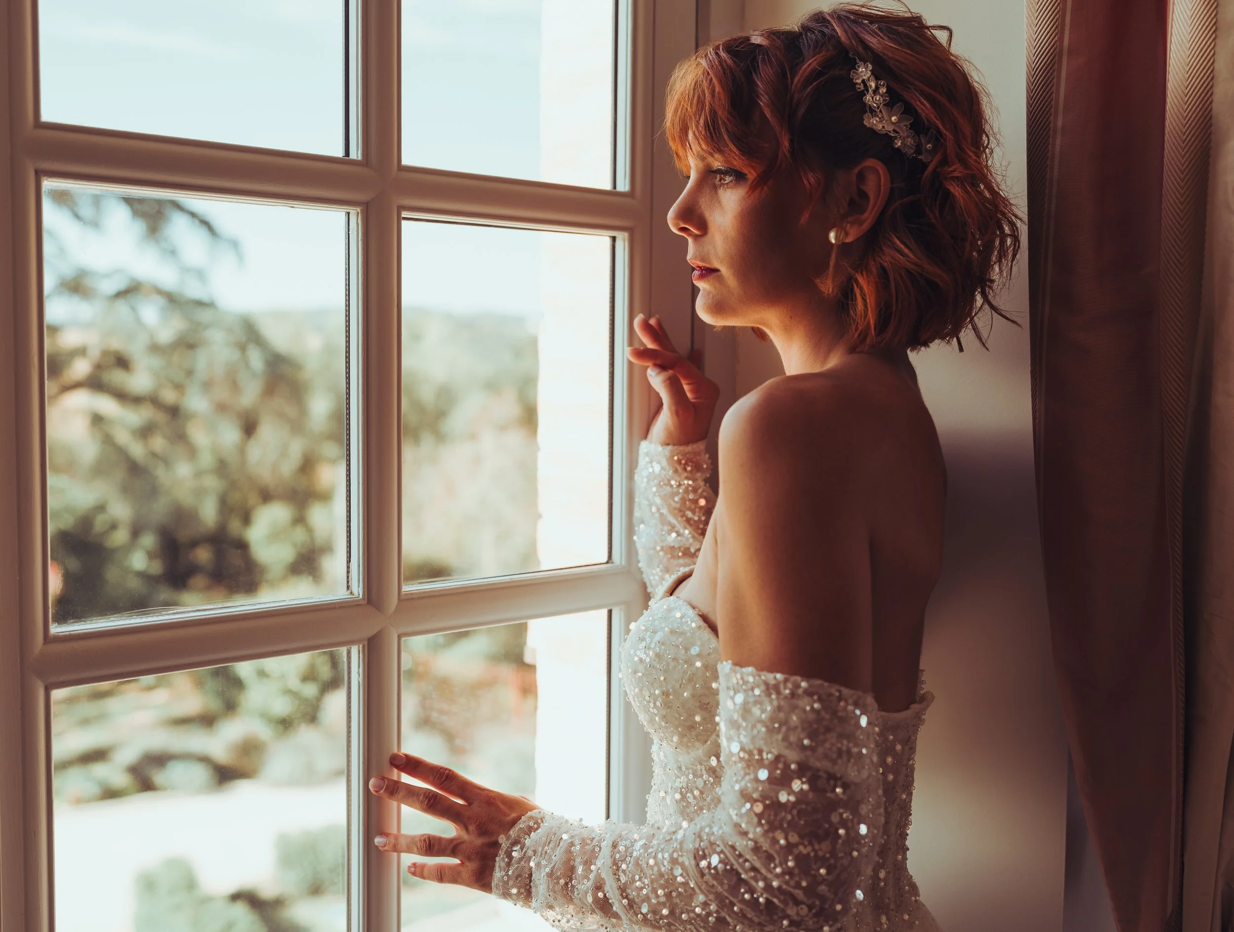 Une mariée regarde par la fenêtre du château de Drudas, portant une robe de mariée en sequin crée par Couture et Mariage à Plaisance du Touch.