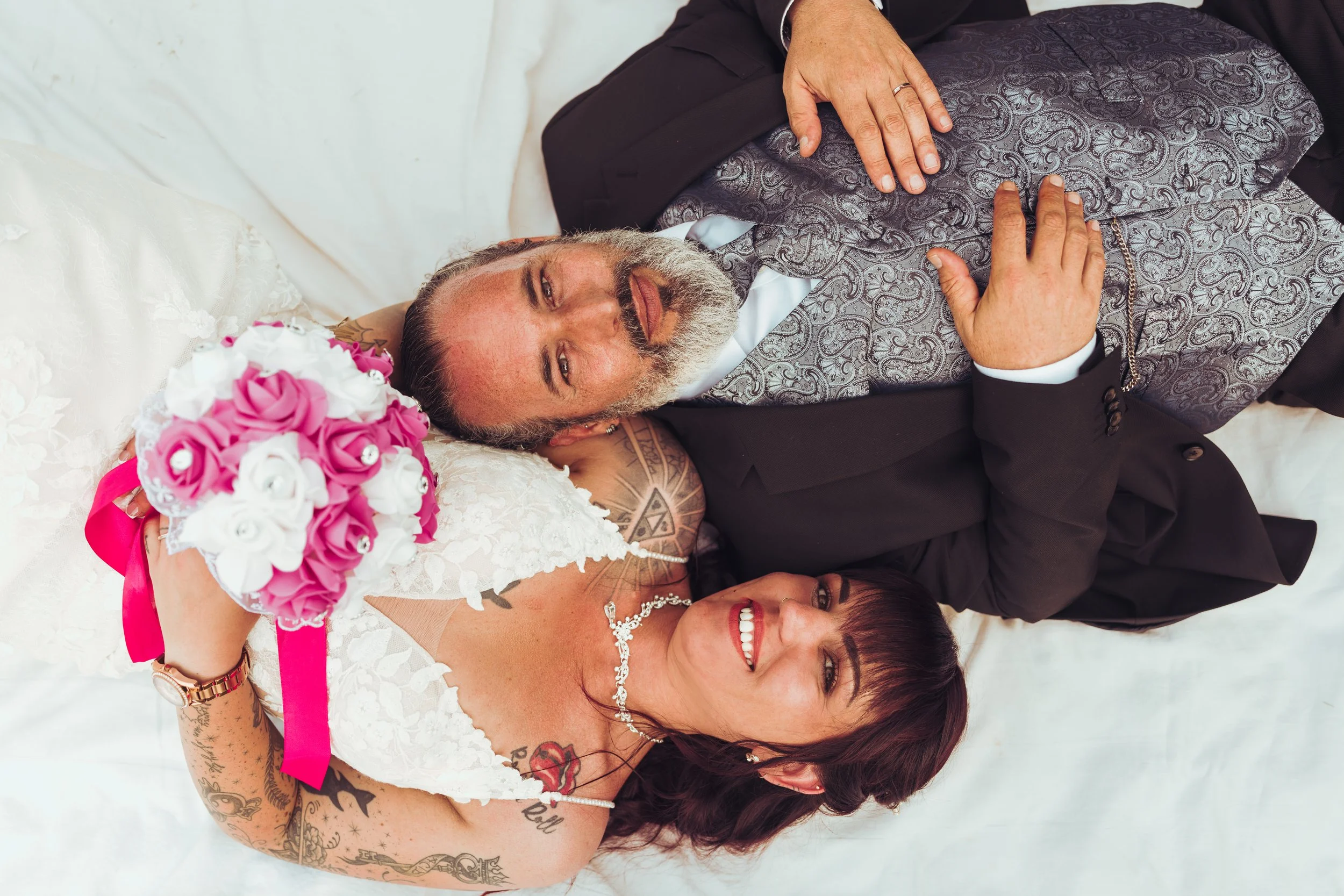 Un couple de mariés allongés sur un lit, souriant, la mariée tenant un bouquet de fleurs roses et blanches, avec des tatouages visibles sur ses bras et une robe blanche pour leur mariage à Auch dans le Gers. 