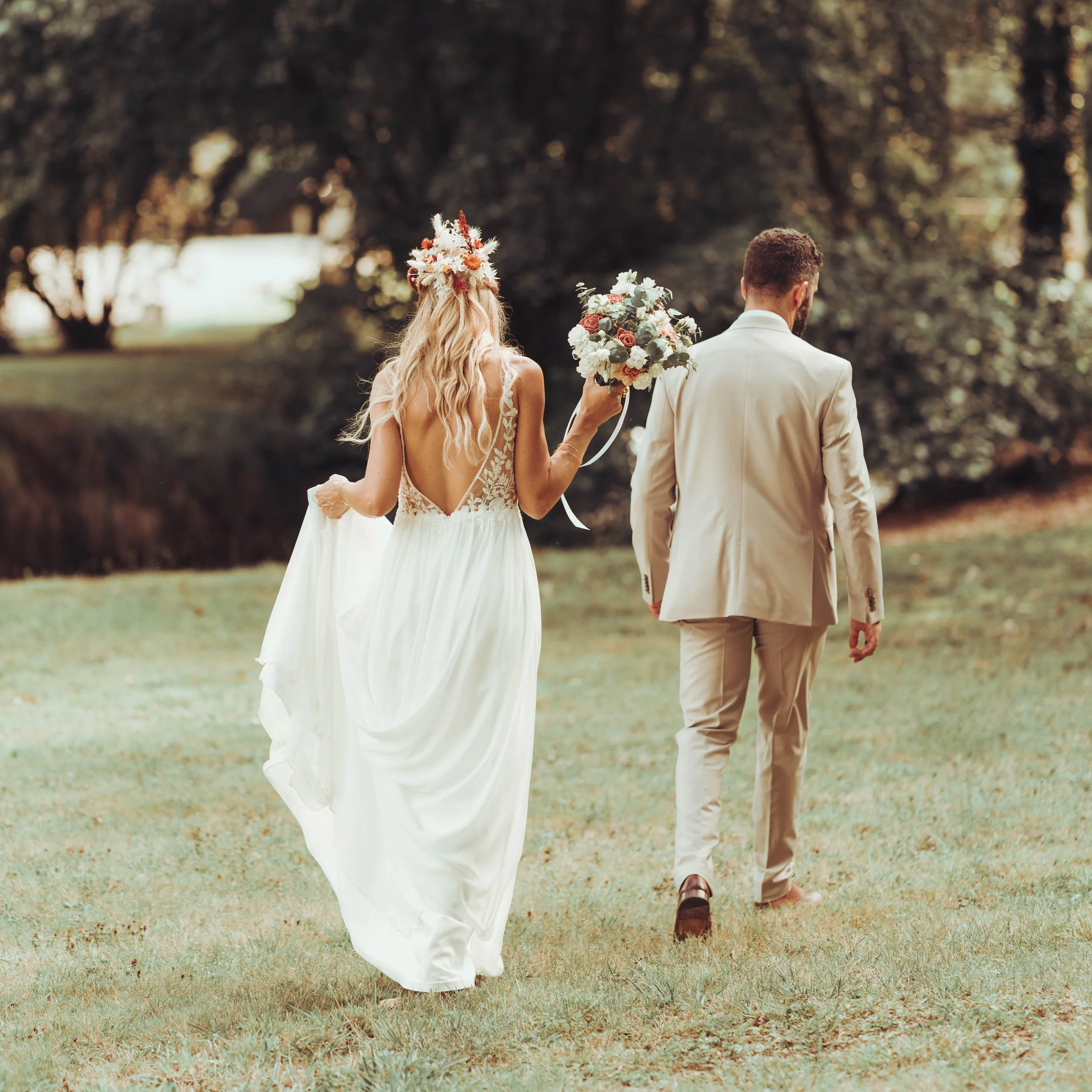 Un couple habillé pour un mariage marche dans le parc du moulin de Rudelle proche de Toulouse avec un arbre en arrière-plan. La femme porte une robe de mariée blanche avec un bouquet, et l'homme porte un costume beige.