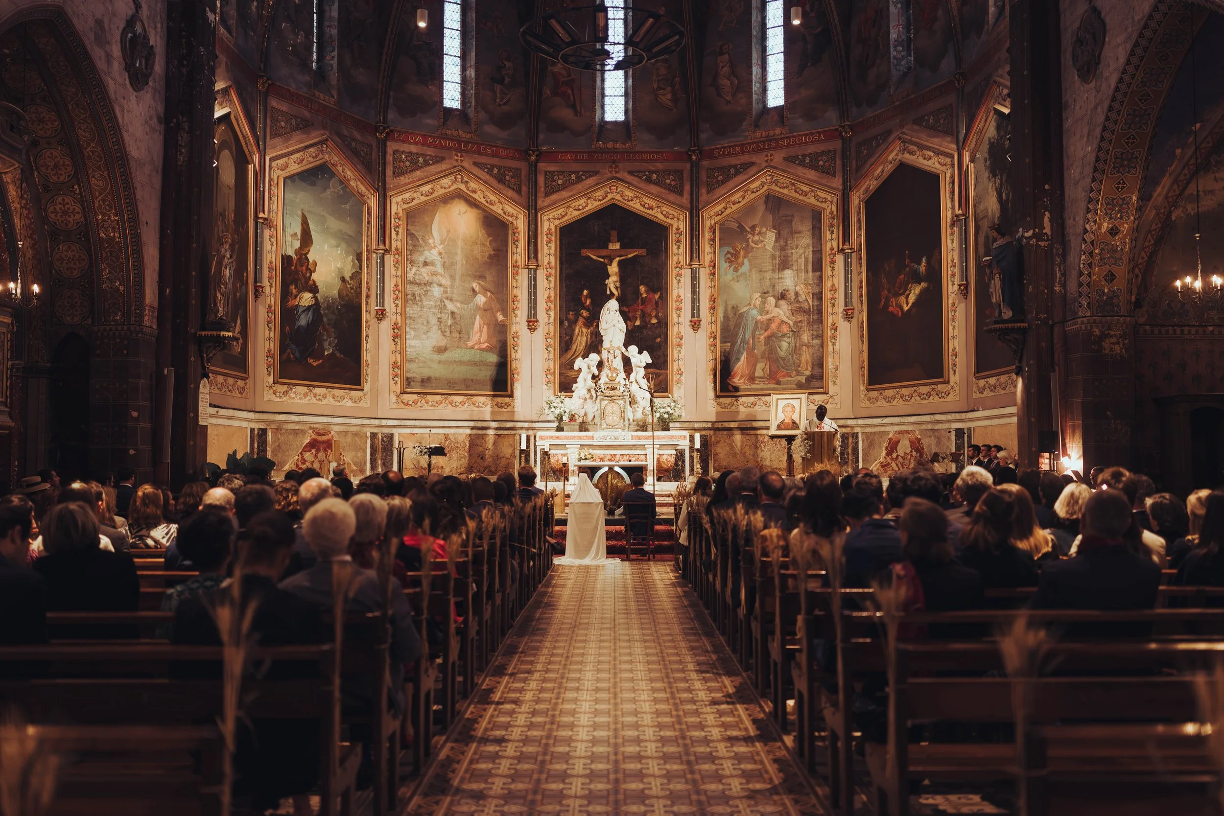 Une cérémonie religieuse de mariage proche de Toulouse dans une ancienne cathédrale avec des dizaines de personnes assises dans la nef, décorée de peintures religieuses et de vitraux, avec une mariée en robe blanche devant l'autel.