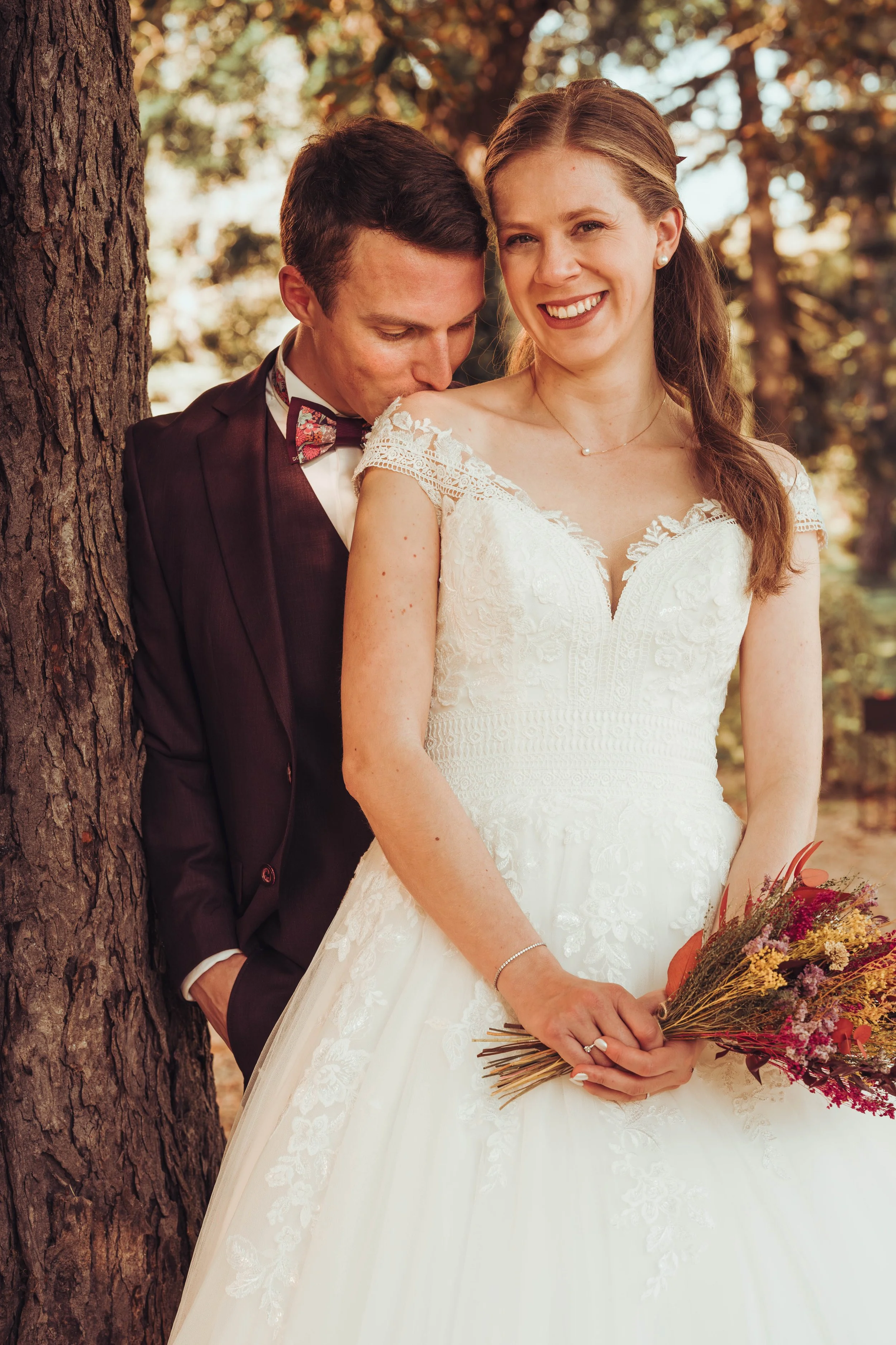 Un couple de mariés pose en plein air au manoir du Thouron proche de Toulouse, avec la mariée tenant un bouquet de fleurs et portant une robe blanche, tandis que l'homme porte un costume foncé avec un nœud papillon à motifs. 