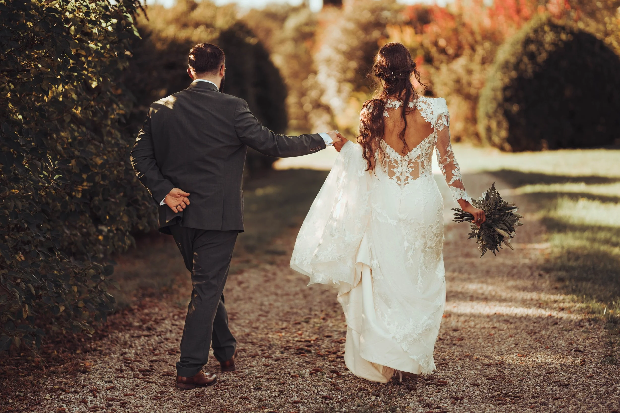 Un couple de jeunes mariés marche dans une forêt en automne au château de Fajac la relenque, la femme en robe de mariée blanche et le homme en costume gris.