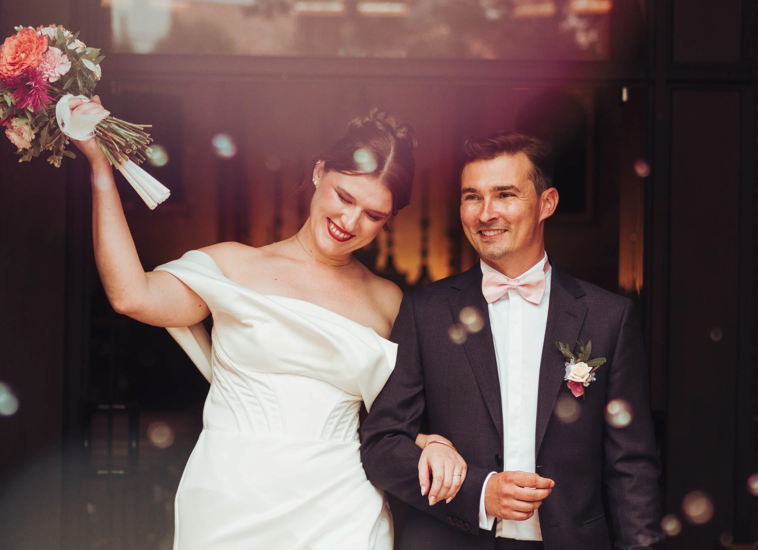 Un couple de mariés souriant, la femme en robe blanche et l'homme en costume noir avec nœud papillon rose, tenant la main, lors d'une cérémonie de mariage religieuse proche de Toulouse, en Occitanie. 