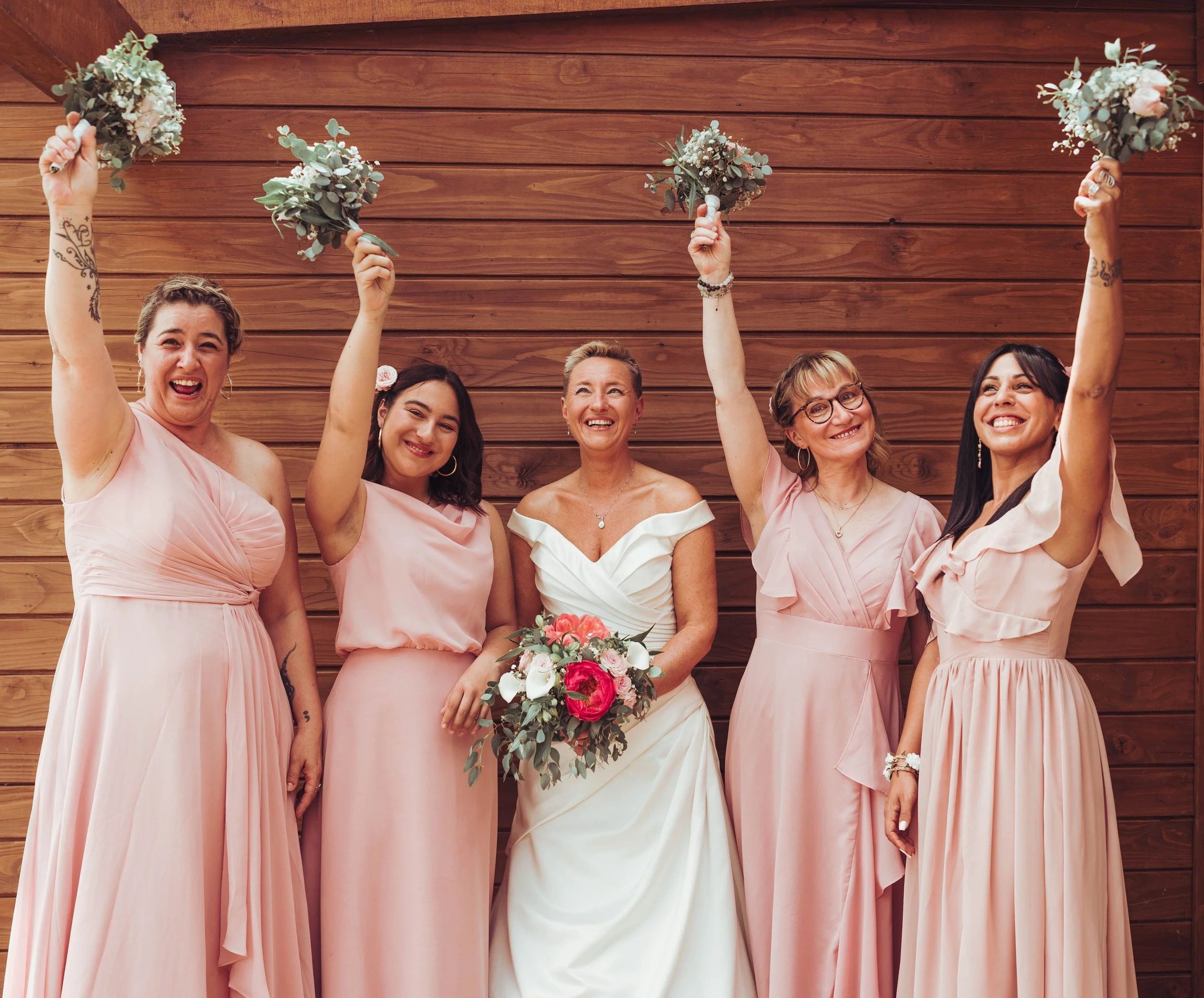 La mariée et ses demoiselles d'honneur proche de Toulouse, lèvent fièrement leurs bouquets de fleurs, posant devant un mur en bois, célébrant un mariage.