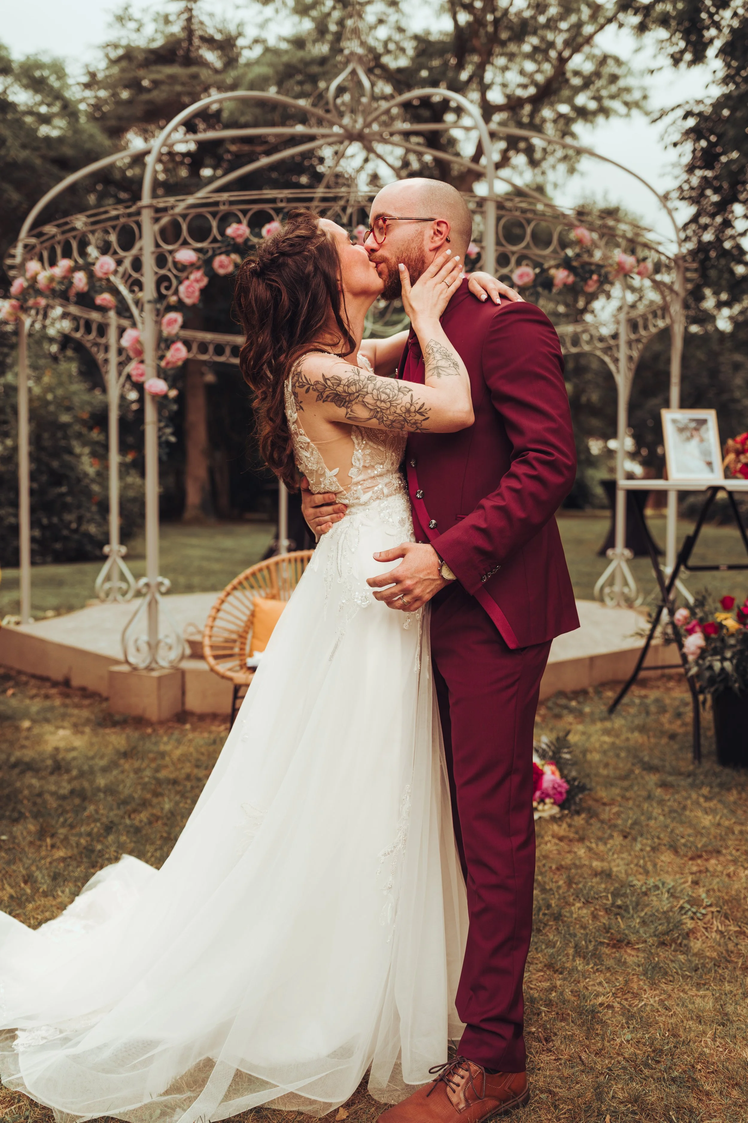 Un couple en robe de mariage et costume partage un baiser lors de leur mariage en plein air au Domaine de Lalanne proche de Toulouse, avec une structure en métal décorée de roses roses en arrière-plan.