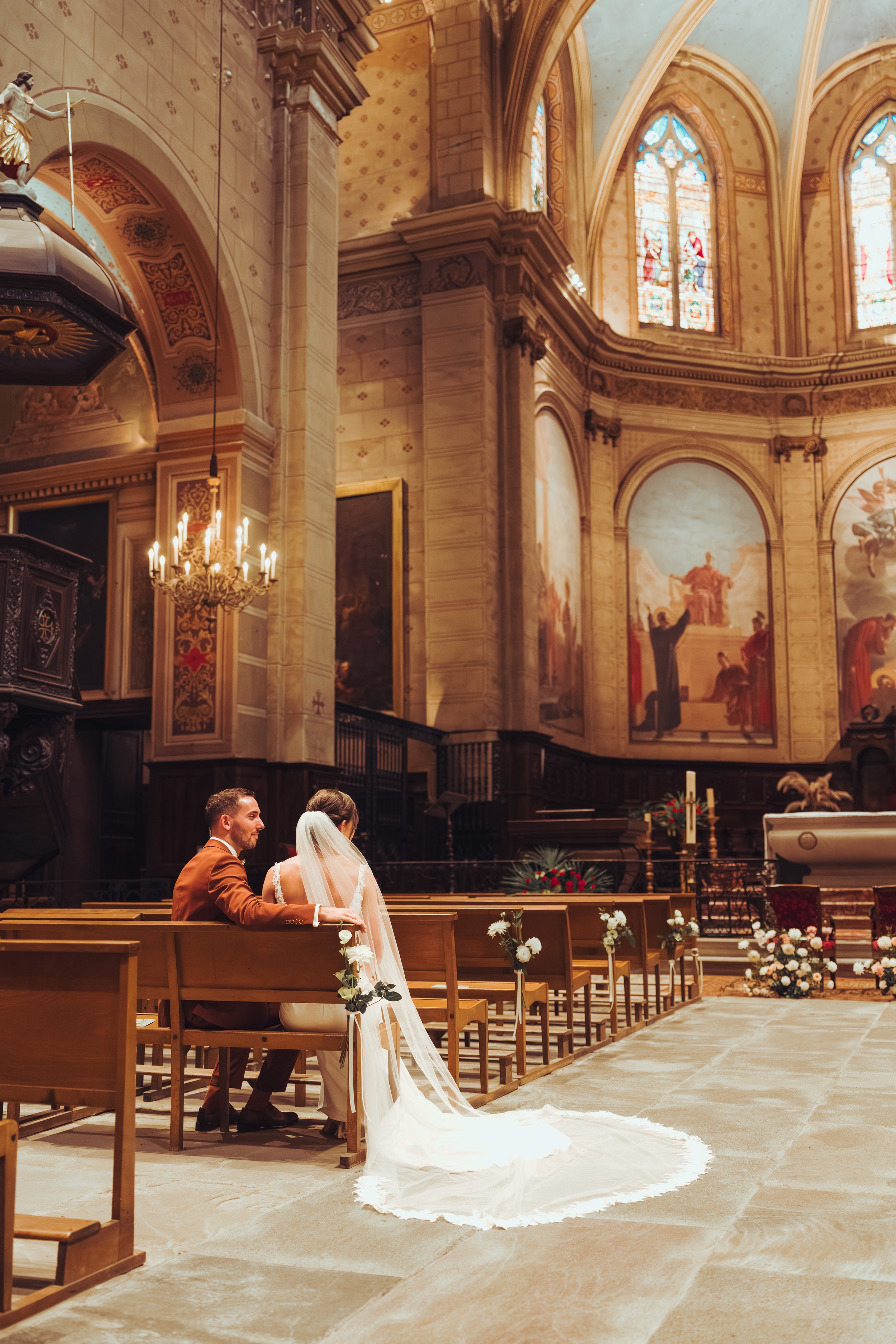 Un couple de mariés assis dans une église gothique après leur cérémonie religieuse avec des vitraux colorés et des fresques, ambiance religieuse et romantique proche de Toulouse. 
