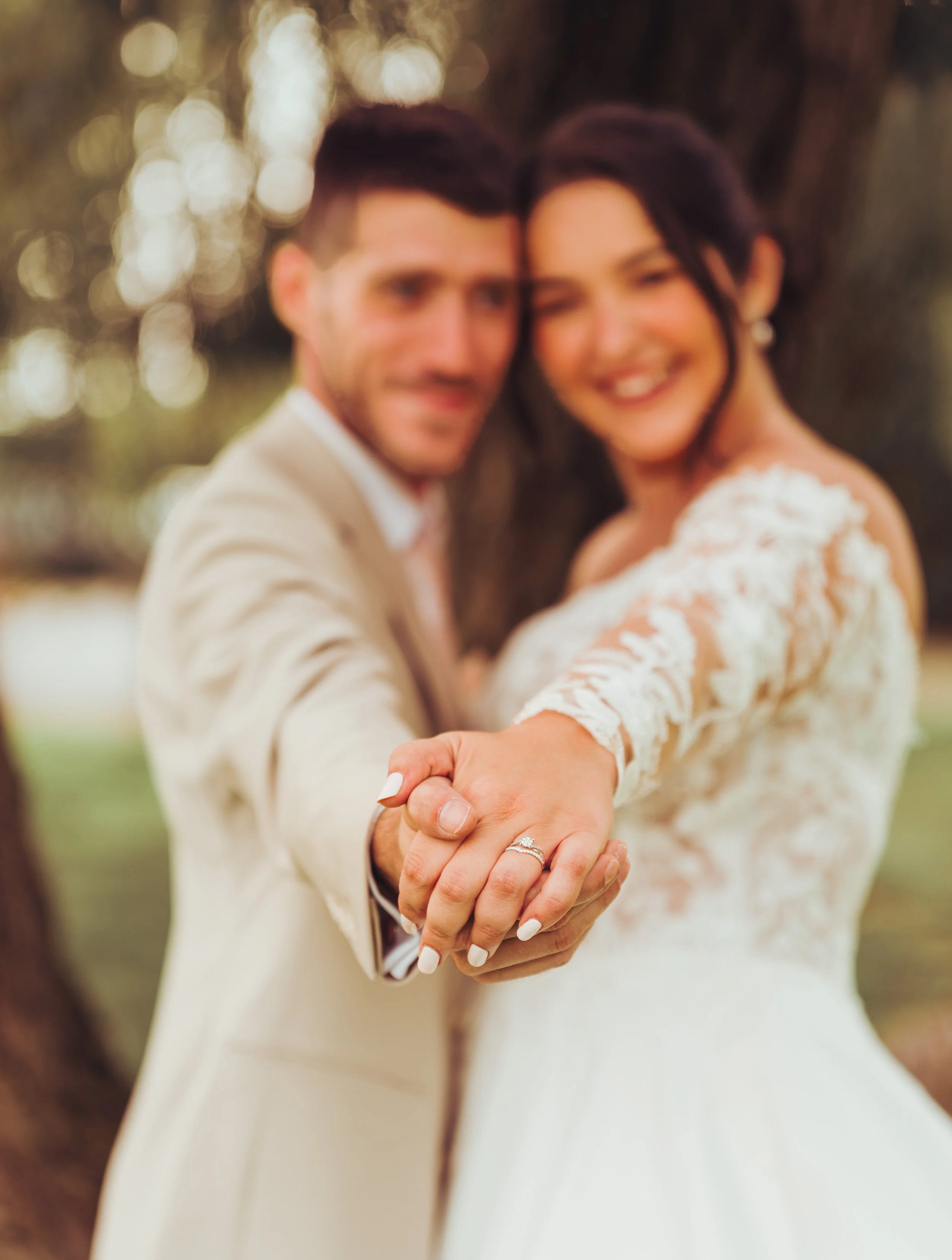 Un couple de mariés souriants au Mas des Canelles près de Toulouse, en Occitanie, tenant la main tendue vers la caméra, avec une alliance de mariage visible, lors d'une séance photo en extérieur après leur cérémonie laïque