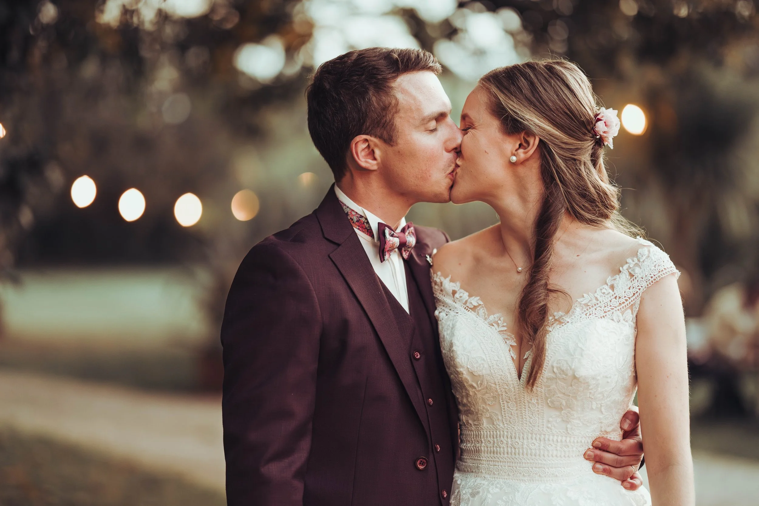 Un couple de mariés est en train de s'embrasser lors d'un mariage en plein air au manoir du Thouron proche de Toulouse, en Occitanie, avec des lumières d'ambiance floues en arrière-plan.