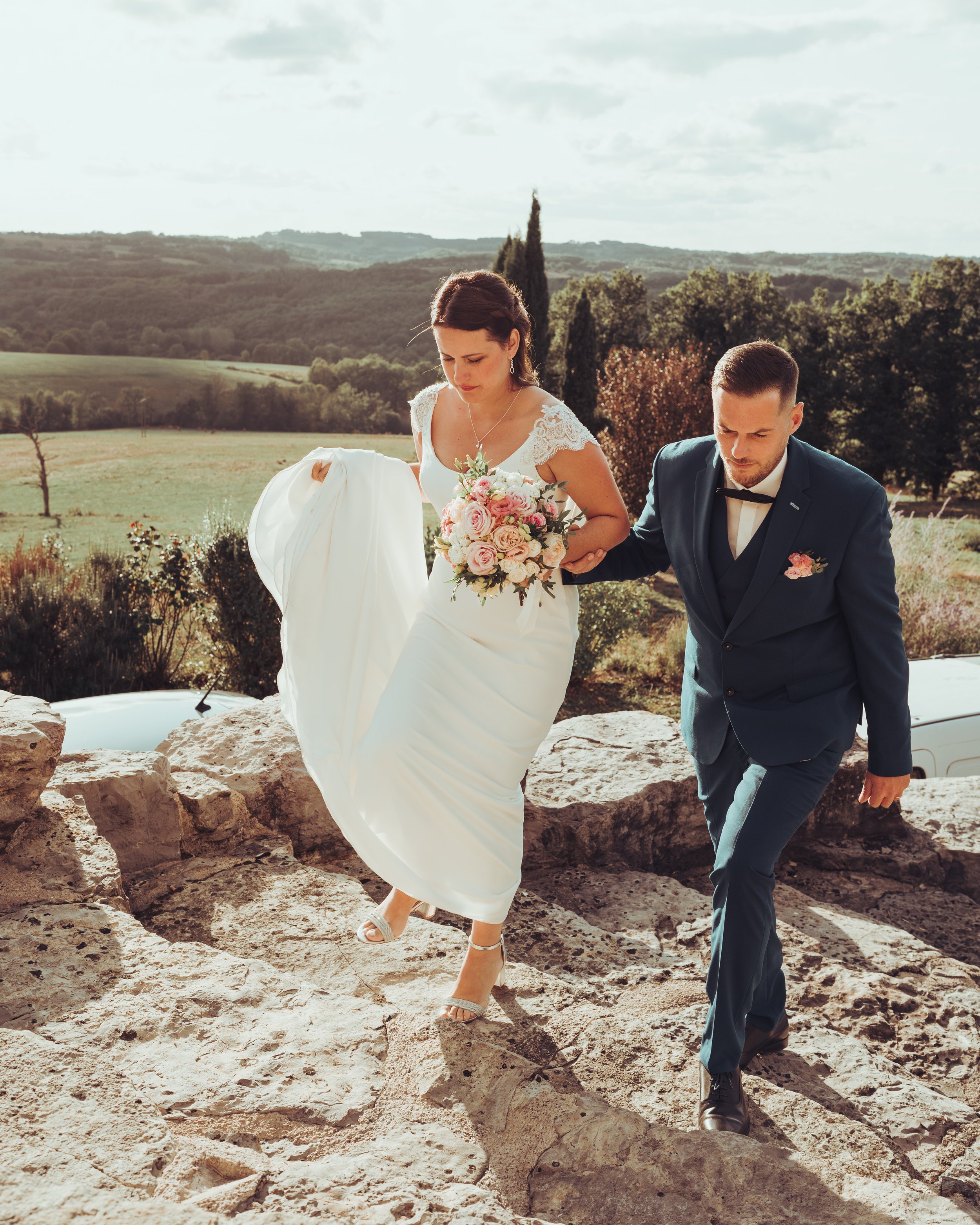 Un couple de mariés marche sur un sentier rocheux en pleine nature dans un décor évoquant la Toscane, la femme porte une robe de mariage blanche et tient un bouquet de fleurs roses, l'homme porte un costume bleu foncé.