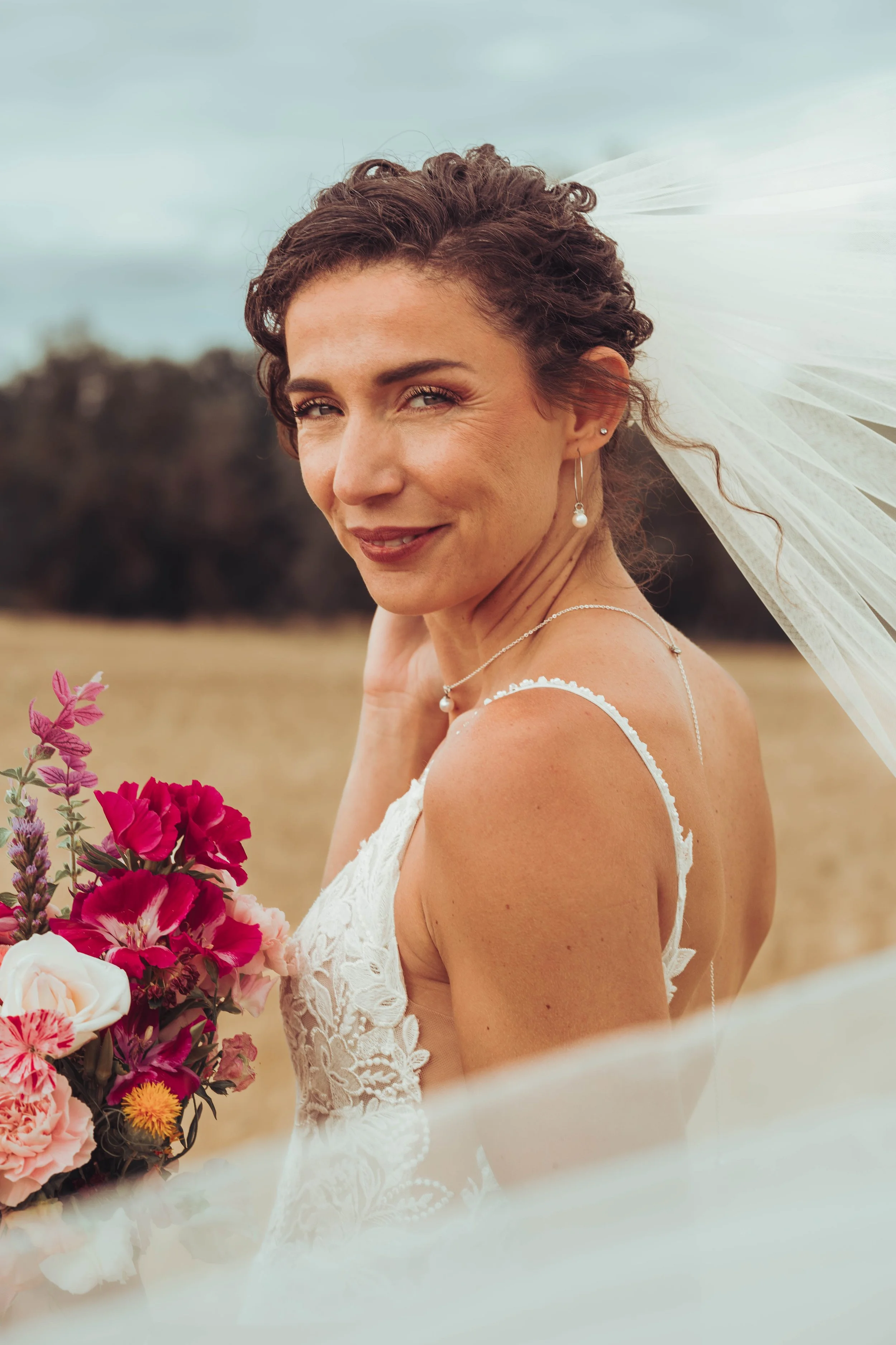 Femme mariée dans un champ proche de Béziers avec un bouquet de fleurs, portant une robe de mariage en dentelle, portant des bijoux avec un voile blanc flottant au vent.