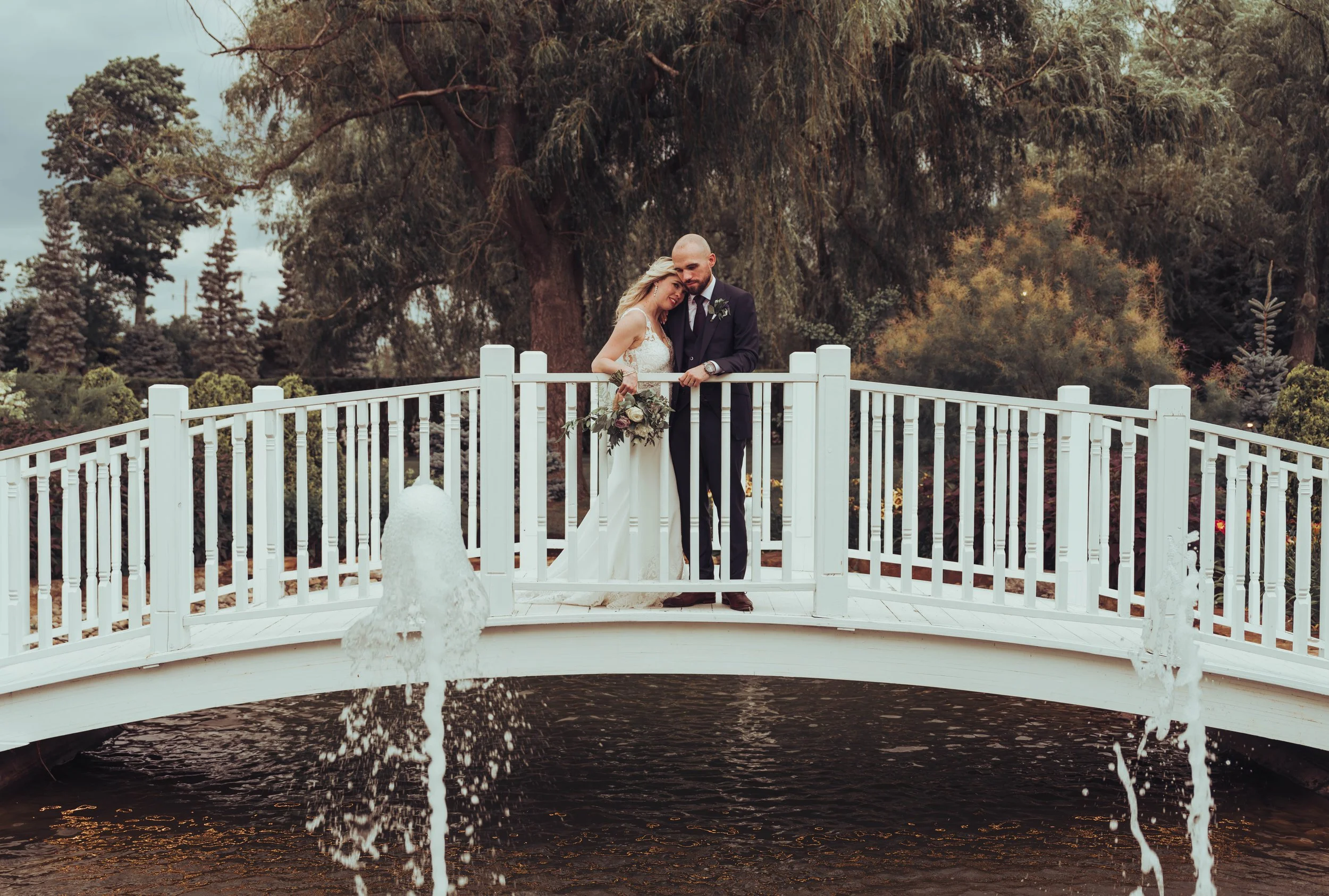 Un couple de mariés se tient sur un pont blanc au-dessus d'une fontaine, entouré d'arbres.