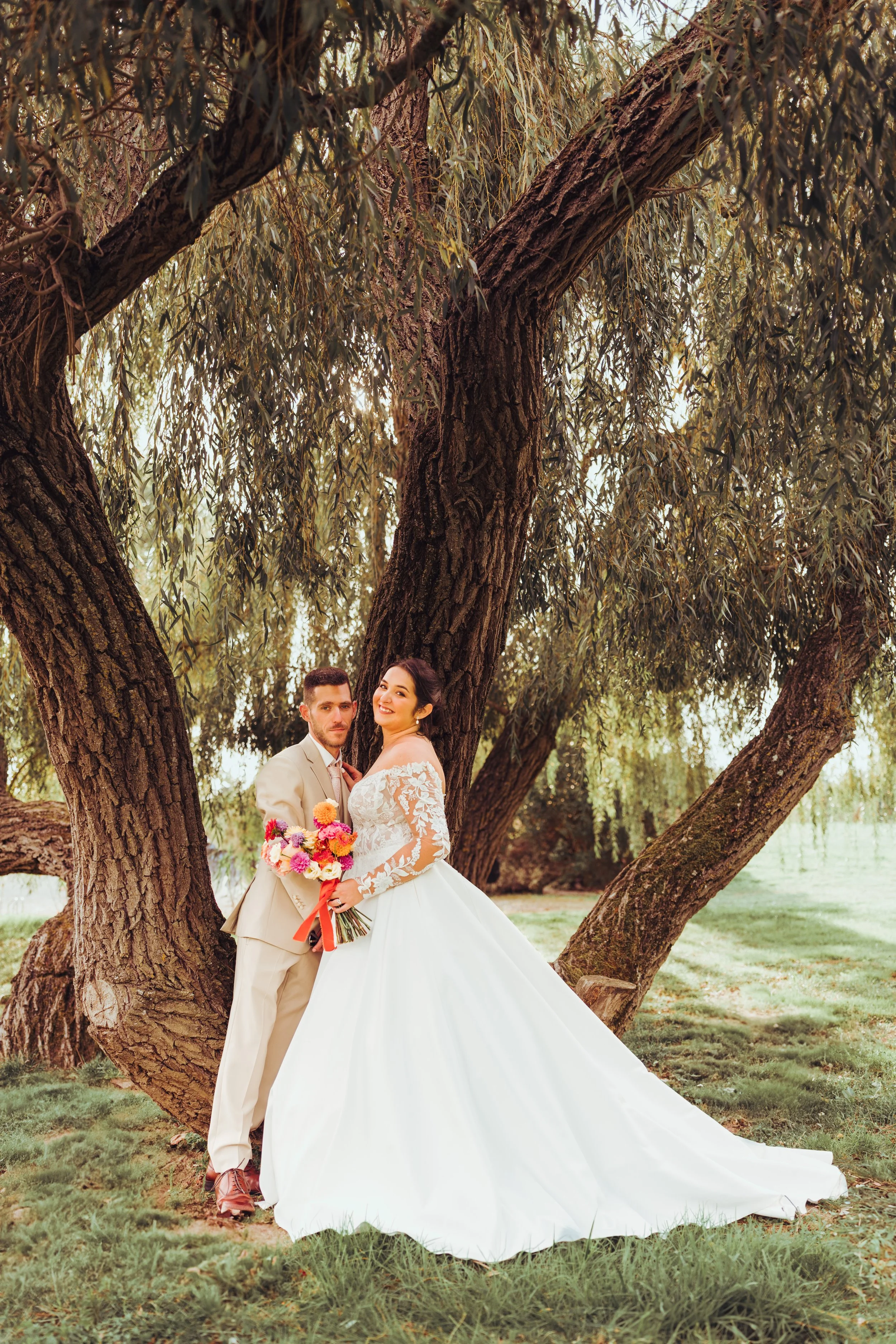 Un couple de mariés posant sous le saule pleureur du Mas des Canelles près de Toulouse, en Occitanie, la femme en robe de mariée blanche et le homme en costume beige, avec un bouquet de fleurs colorées.