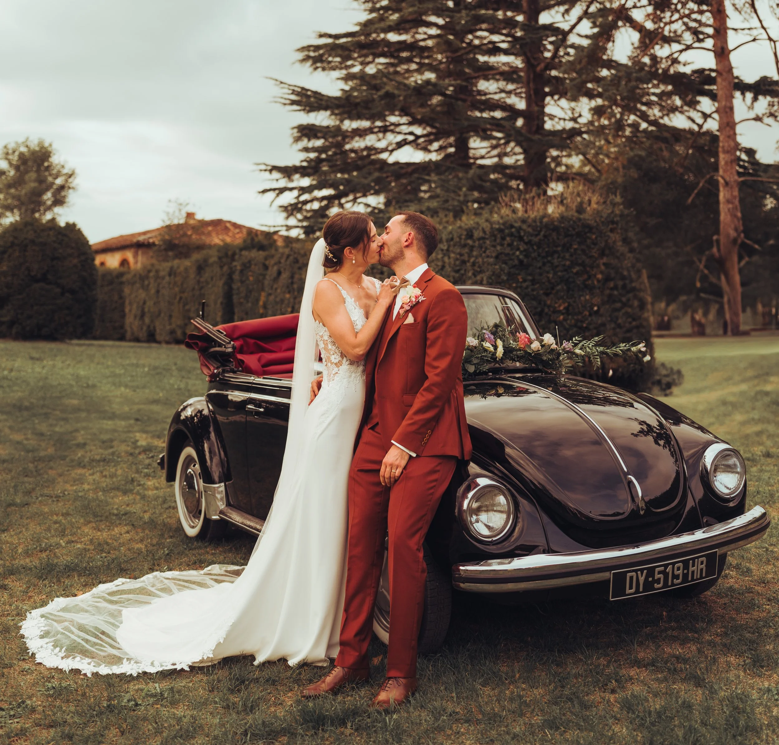 Un couple de mariés en costume et robe de mariage s'embrassent devant une voiture ancienne décorée de fleurs dans un jardin herbeux avec des grands arbres en arrière-plan au chateau de Fajac la relenque