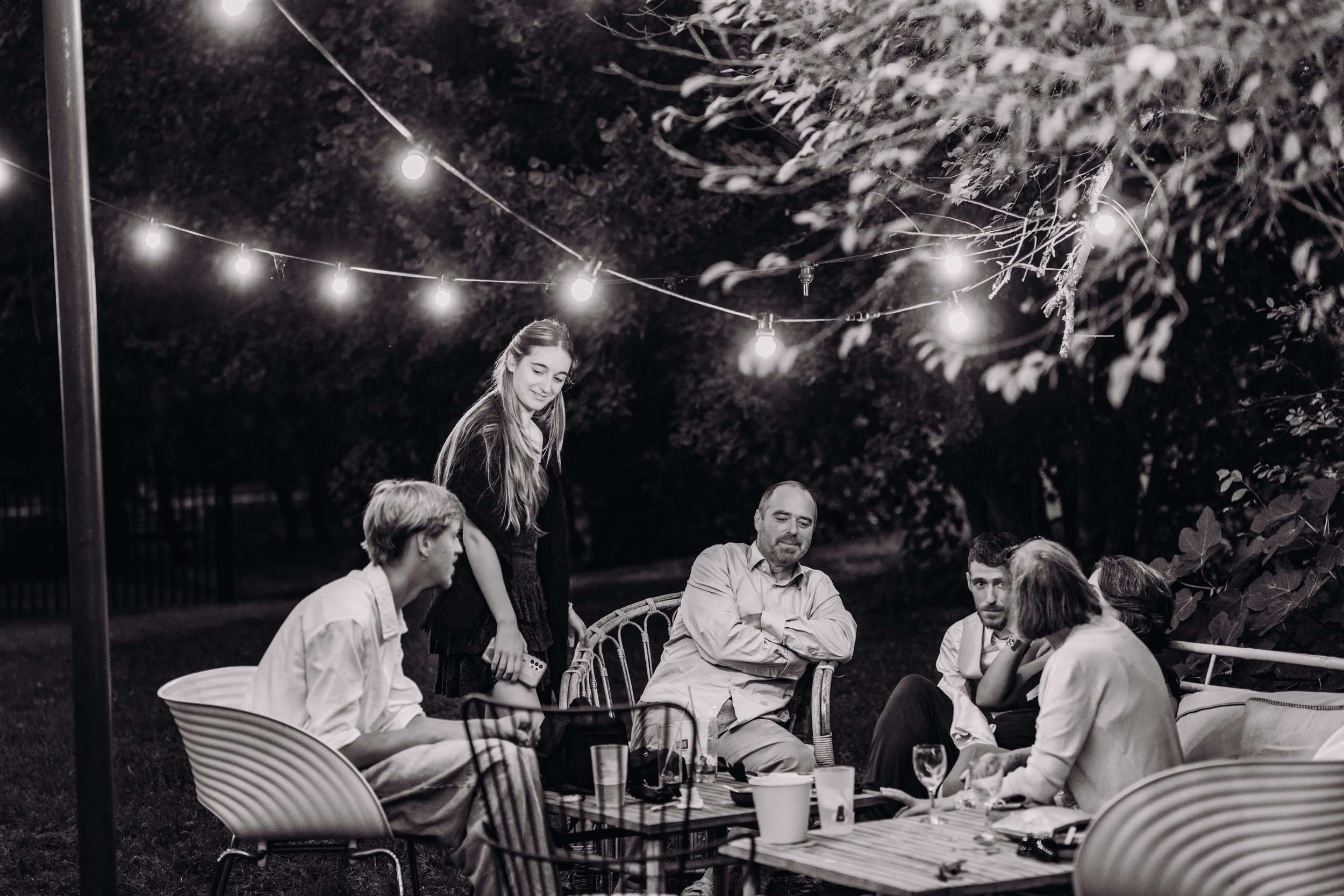 Groupe de sept personnes réunies autour d'une table en plein air au Mas des canelles, proche de toulouse, lors d'une soirée de mariage, sous des guirlandes lumineuses, dans un environnement nocturne avec des arbres en arrière-plan.