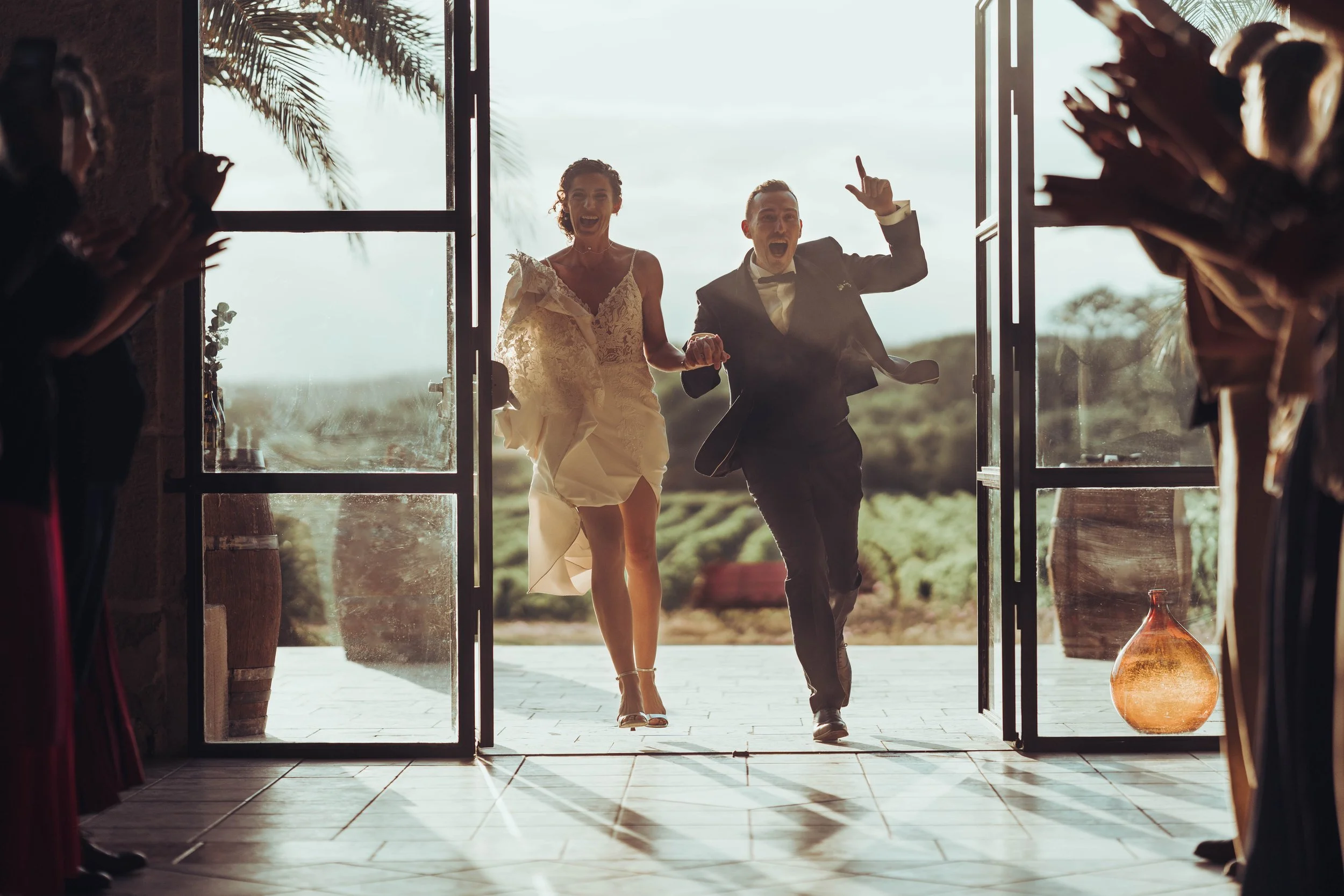 Un couple en vêtements de mariage court et tuxedo courant vers leur sortie lors d'un mariage, sous un ciel ensoleillé, entouré par des invités, avec un décor naturel en arrière-plan.