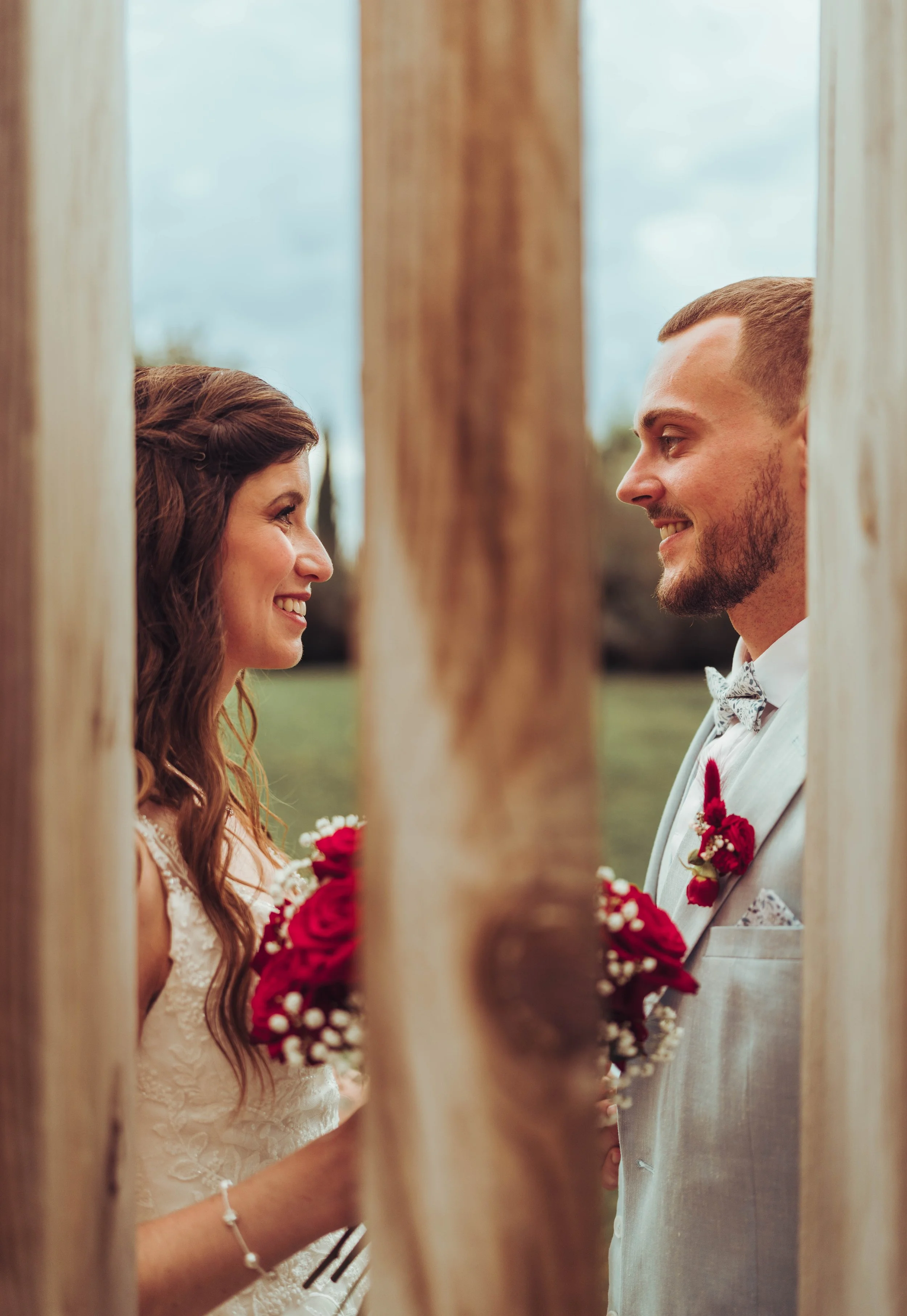 Un couple marié regarde l'un l'autre à travers un cadre en bois, tenant des bouquets de roses rouges, lors de leur mariage au Mas Tolosa.