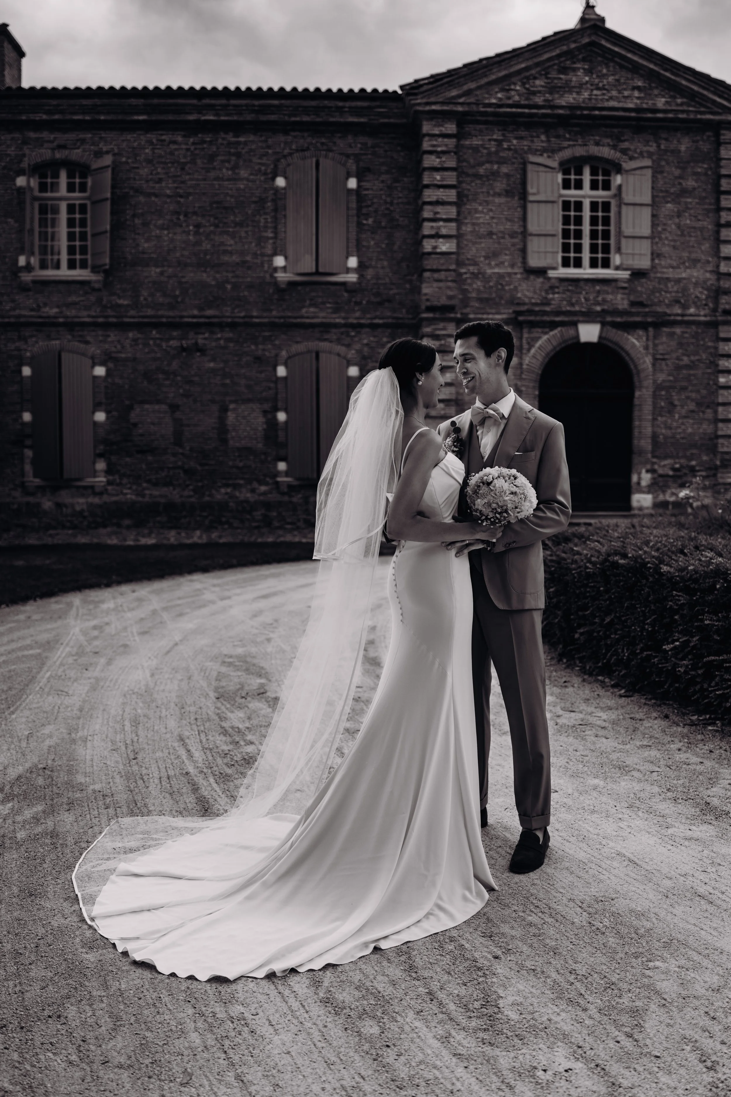 Un couple marié se regarde dans les yeux, échangeant un sourire, devant l'Orangerie des demoiselles (domaine de mariage proche de Toulouse) lors de leur mariage estival