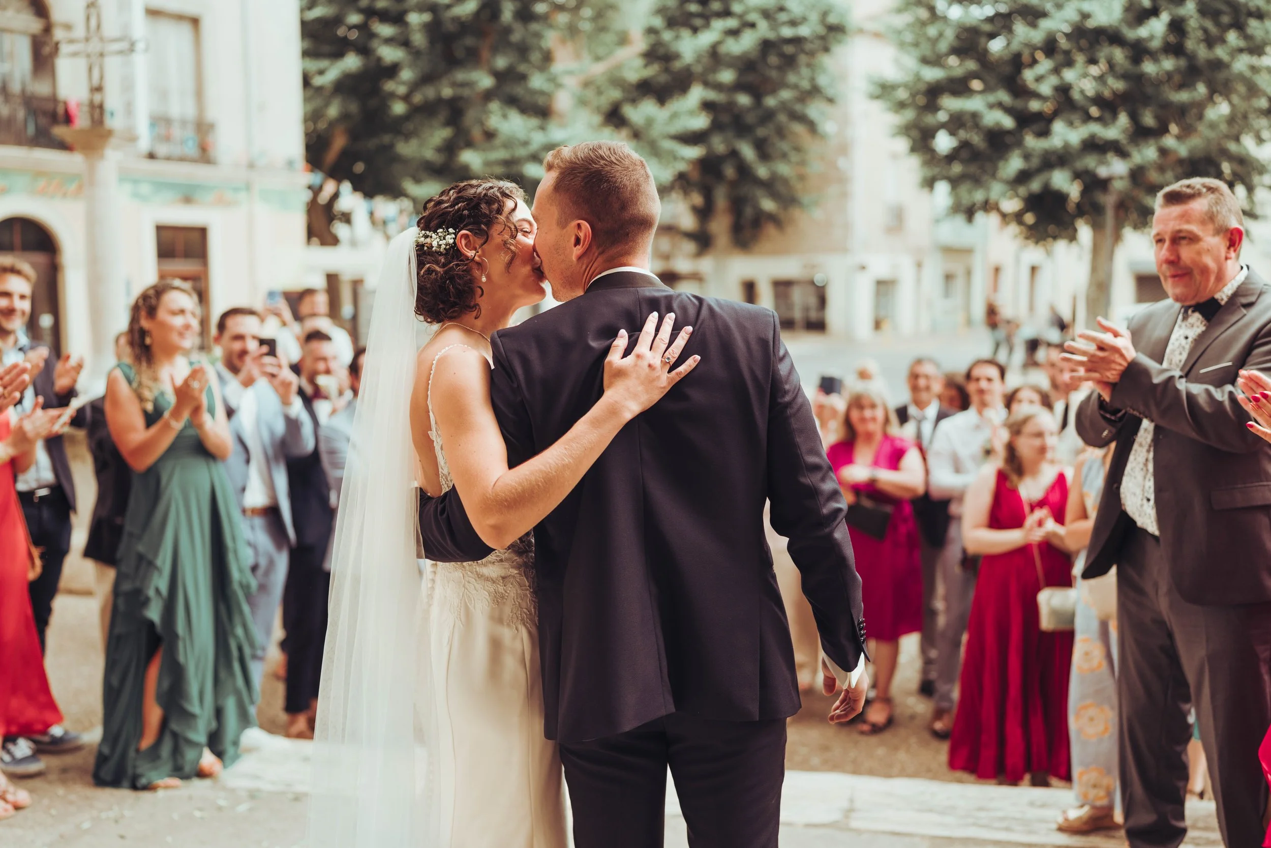 Un couple de mariés s'embrassant à la sortie de leur cérémonie religieuse proche de Béziers lors de leur mariage en plein air, entourés d'invités applaudissant et prenant des photos, en face d'une vieille bâtisse et d'arbres.