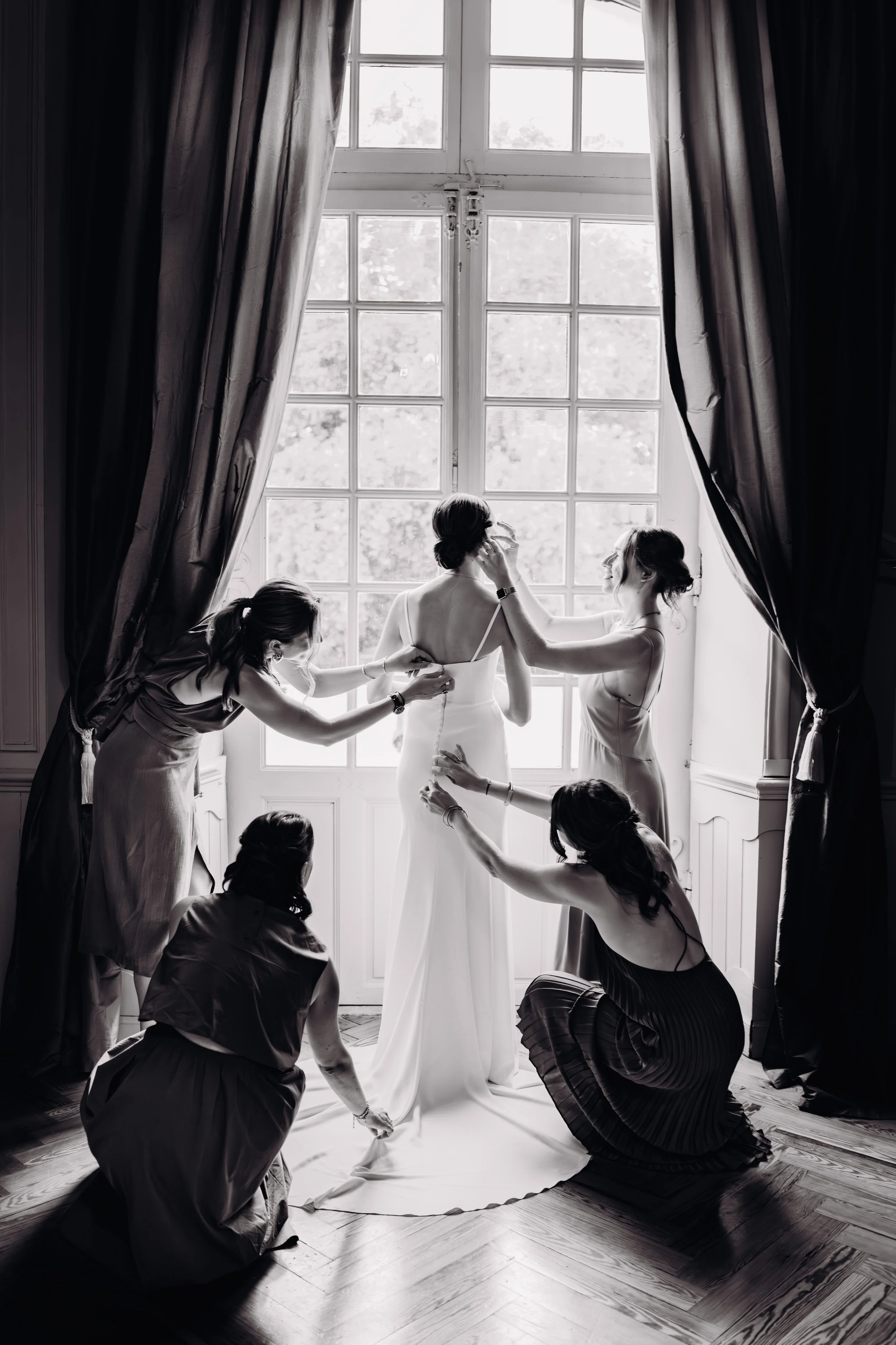 Cinq femmes, probablement des amies ou la famille, assistent à la mariée pendant qu'elle se prépare pour son mariage, en ajustant sa robe près d'une grande fenêtre de l'Orangerie des Demoiselles près de Toulouse, en Occitanie. 