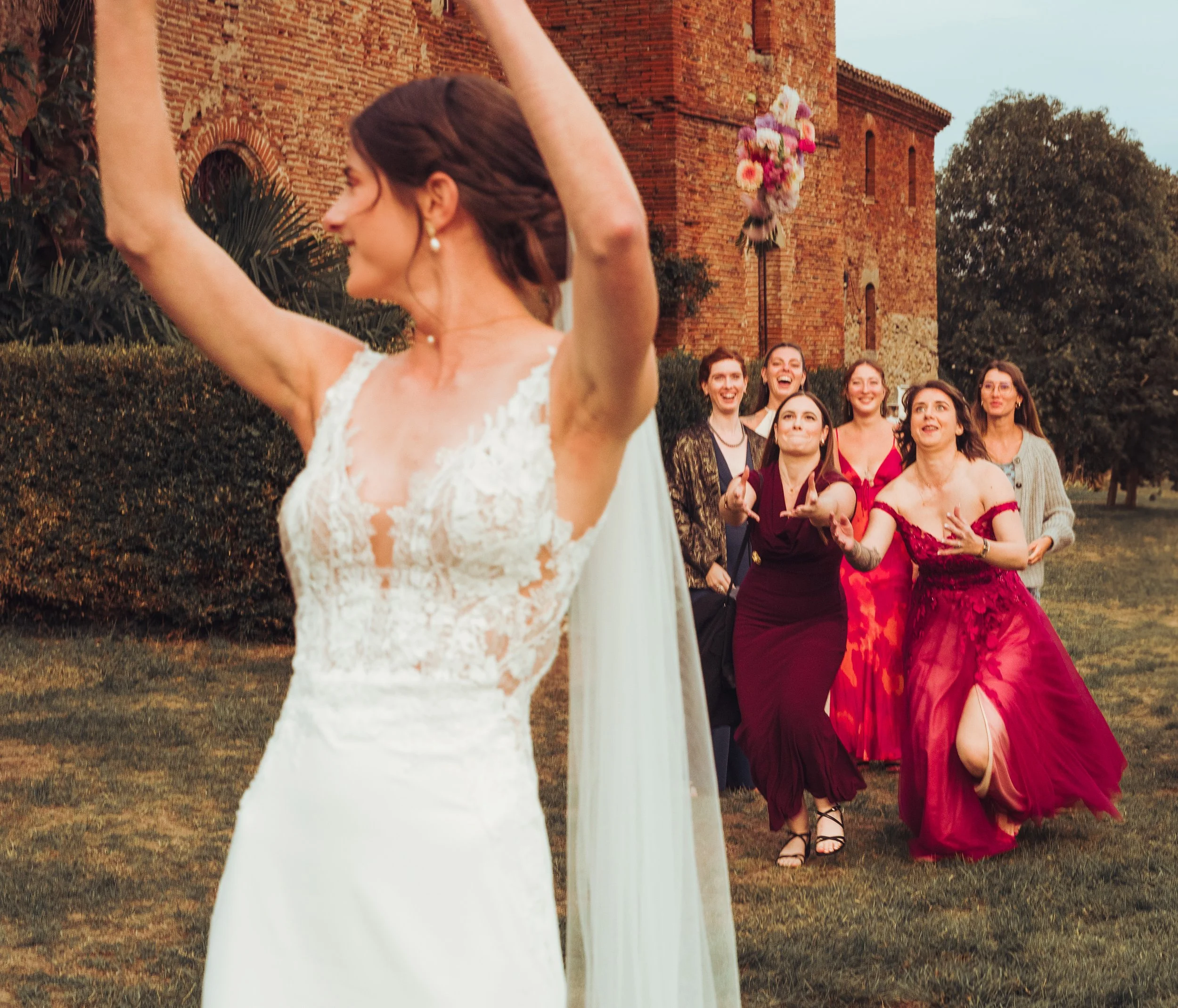 Une mariée en robe blanche, de dos, lançant un bouquet de fleurs vers un groupe de femmes souriantes qui attendent pour le rattraper, avec une vieille église en pierre en arrière-plan au chateau de Fajac la relenque proche de Toulouse. 