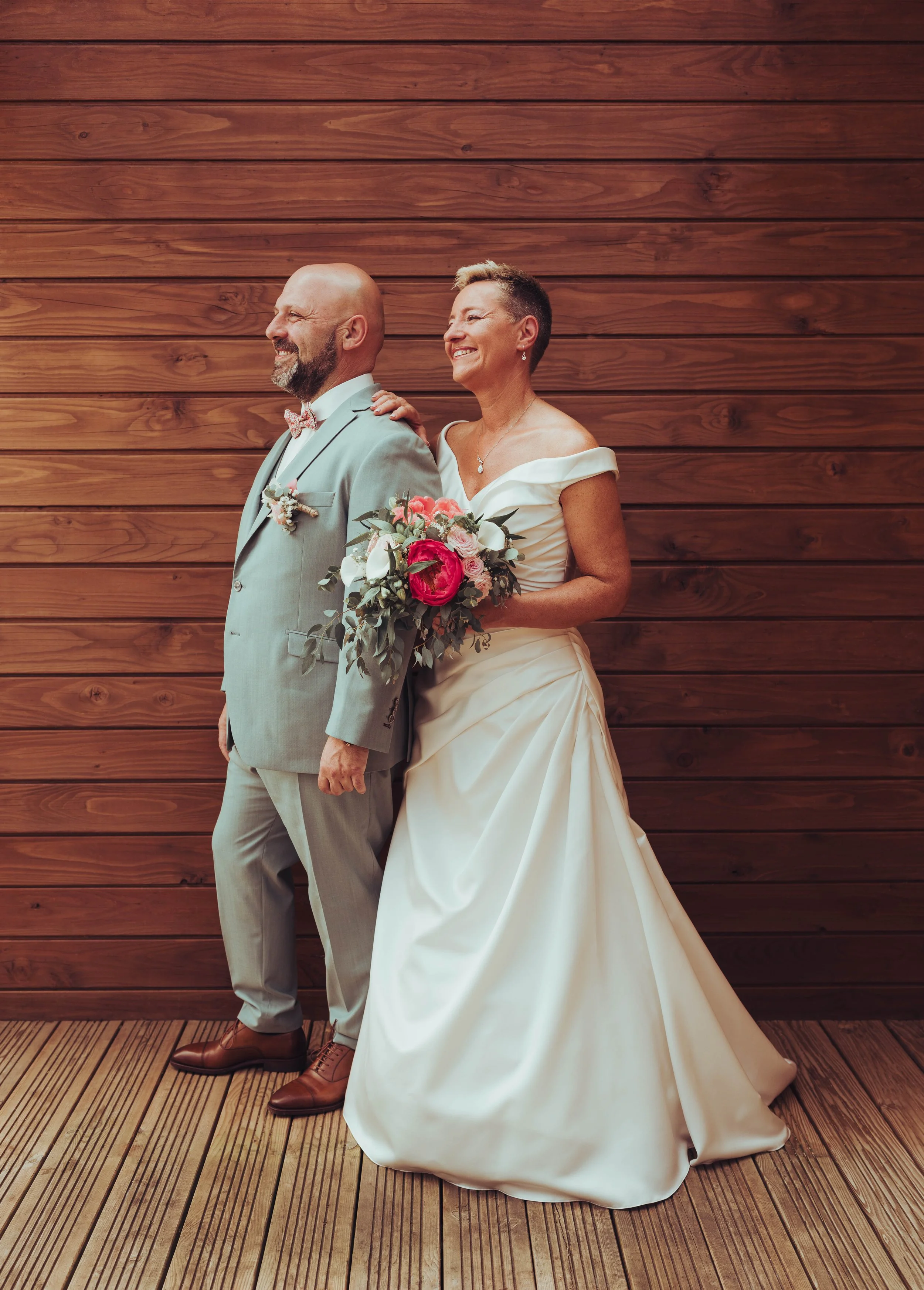 Un couple en mariage proche de Toulouse, souriant, devant un mur en bois. La mariée porte une robe blanche et tient un bouquet de fleurs, tandis que le marié porte un costume gris avec une nœud papillon.