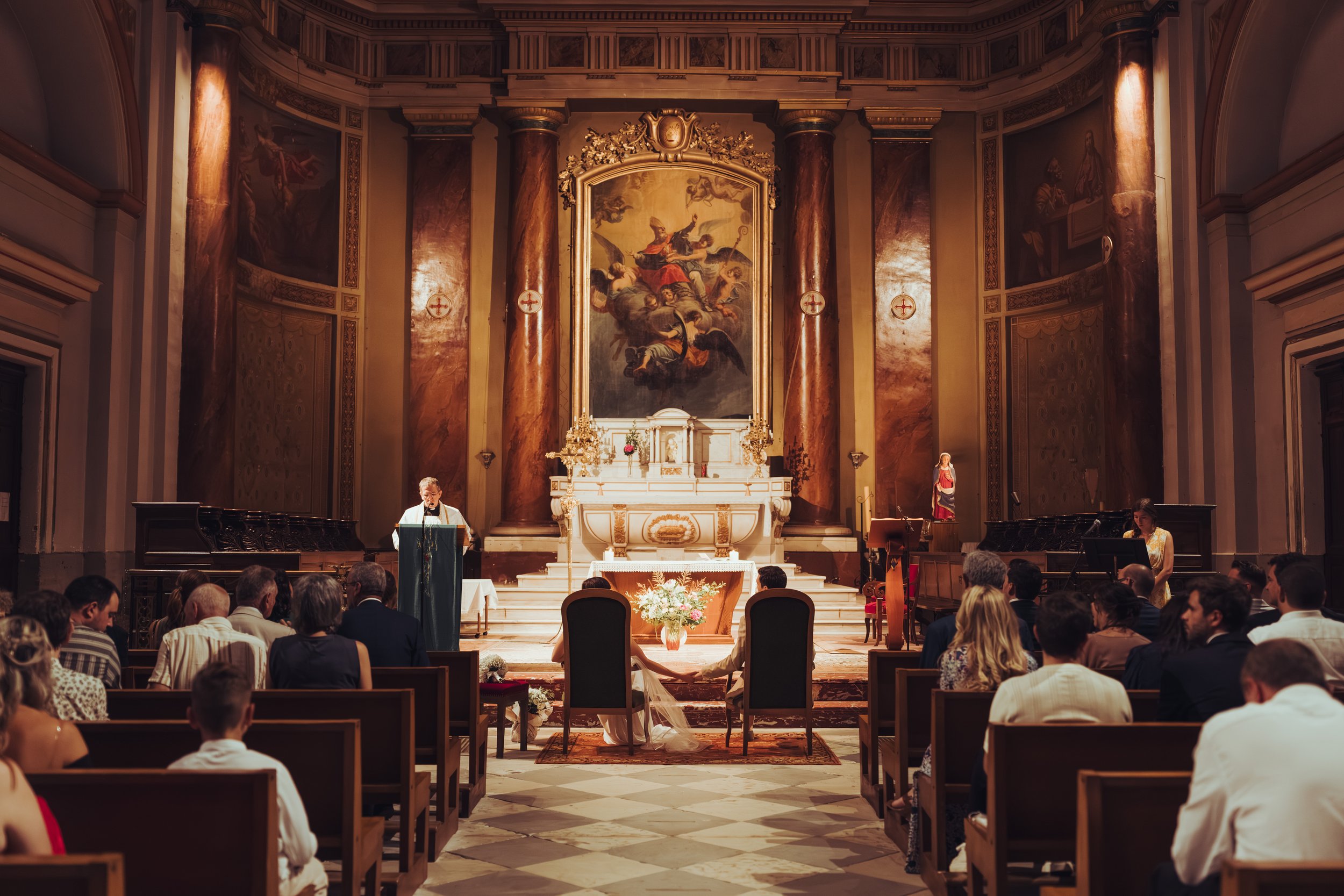 Mariage dans une église avec l'autel au centre, des invités assis et les mariés assis devant eux. Un prêtre ou officiant parle à la cérémonie. Décorations florales et grande peinture religieuse en arrière-plan.