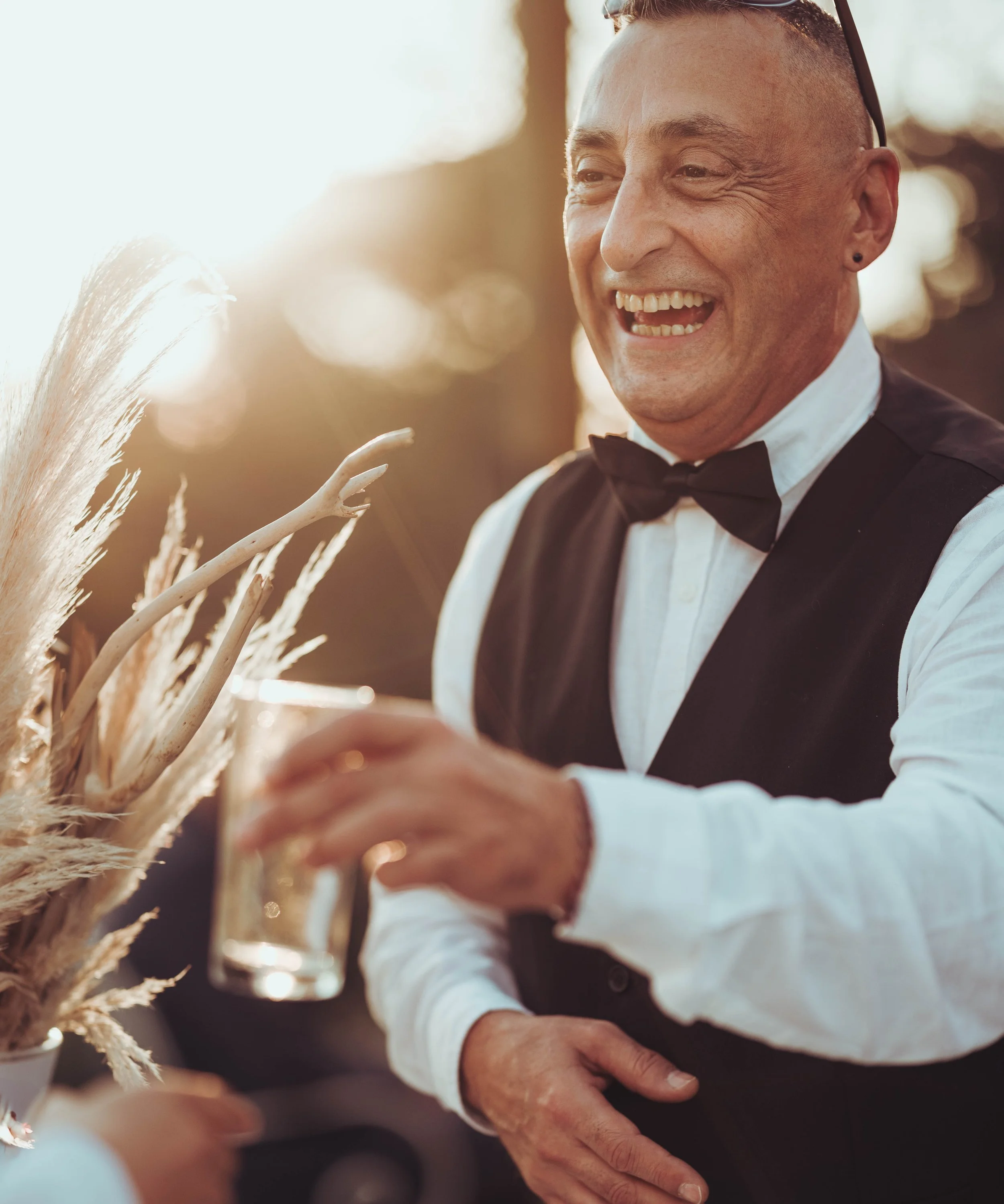 Homme souriant en smoking noir et nœud papillon, tenant un verre à cocktail lors d'une réception en plein air au coucher du soleil au château de Fajac la relenque proche de Toulouse.