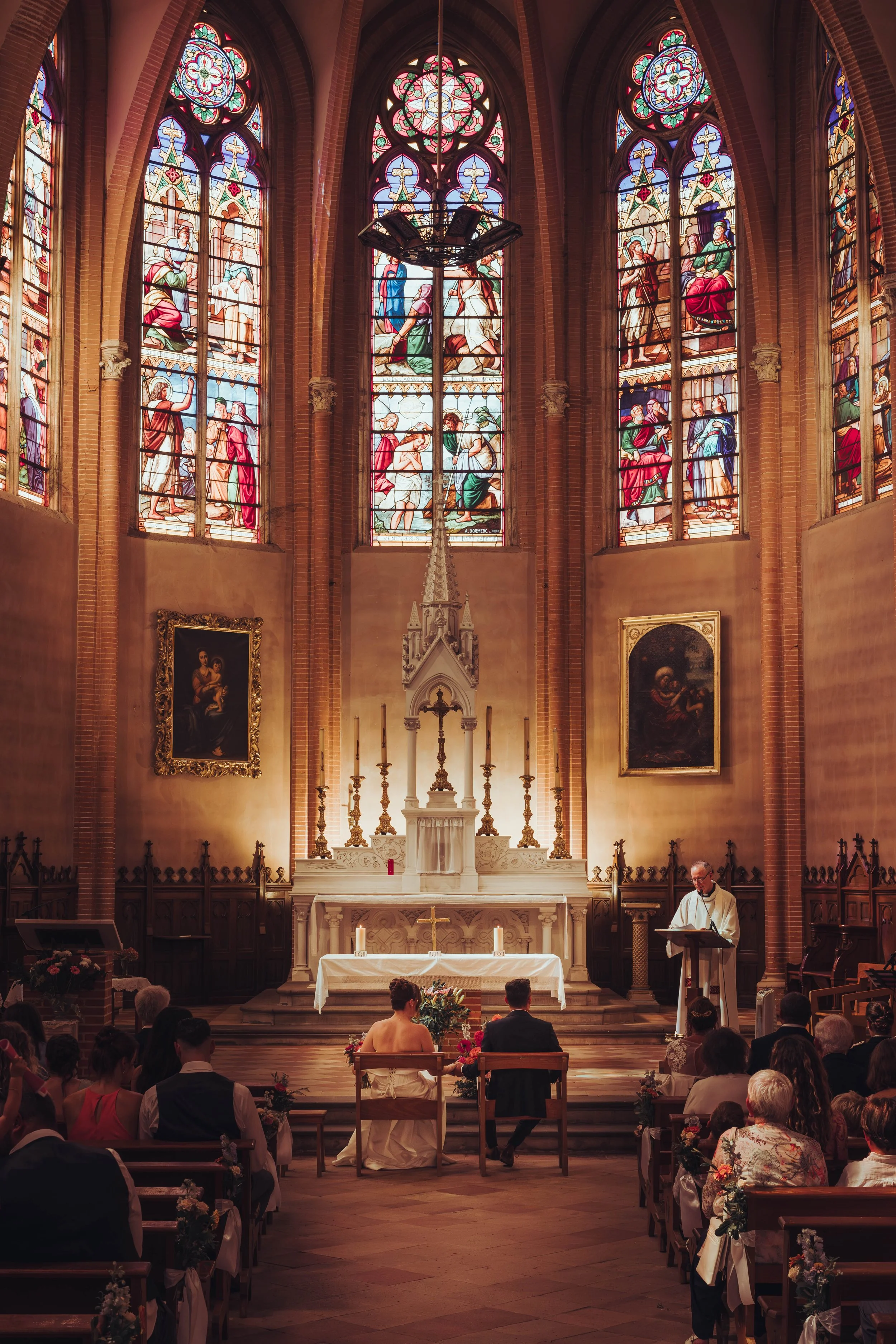 Une cérémonie de mariage dans une église proche Toulouse avec vitraux colorés, un couple devant l'autel, un prêtre à droite, et des invités assis dans la salle.