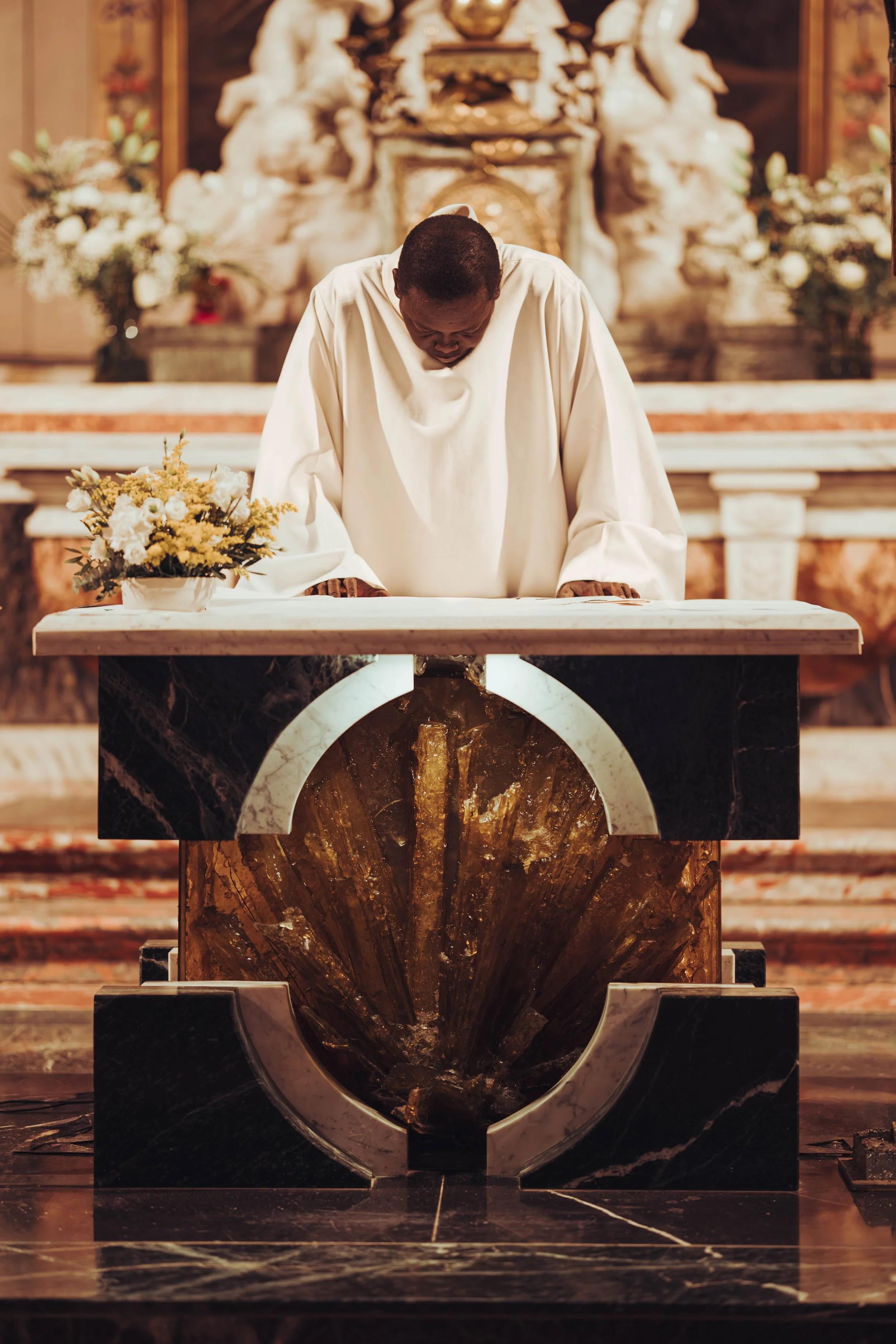 Un prêtre en habits blancs prie dans une église proche de Toulouse, en Occitanie  devant un autel en marbre avec une fontaine ou un symbole en pierre, décoré de fleurs colorées sur le côté juste avant un mariage