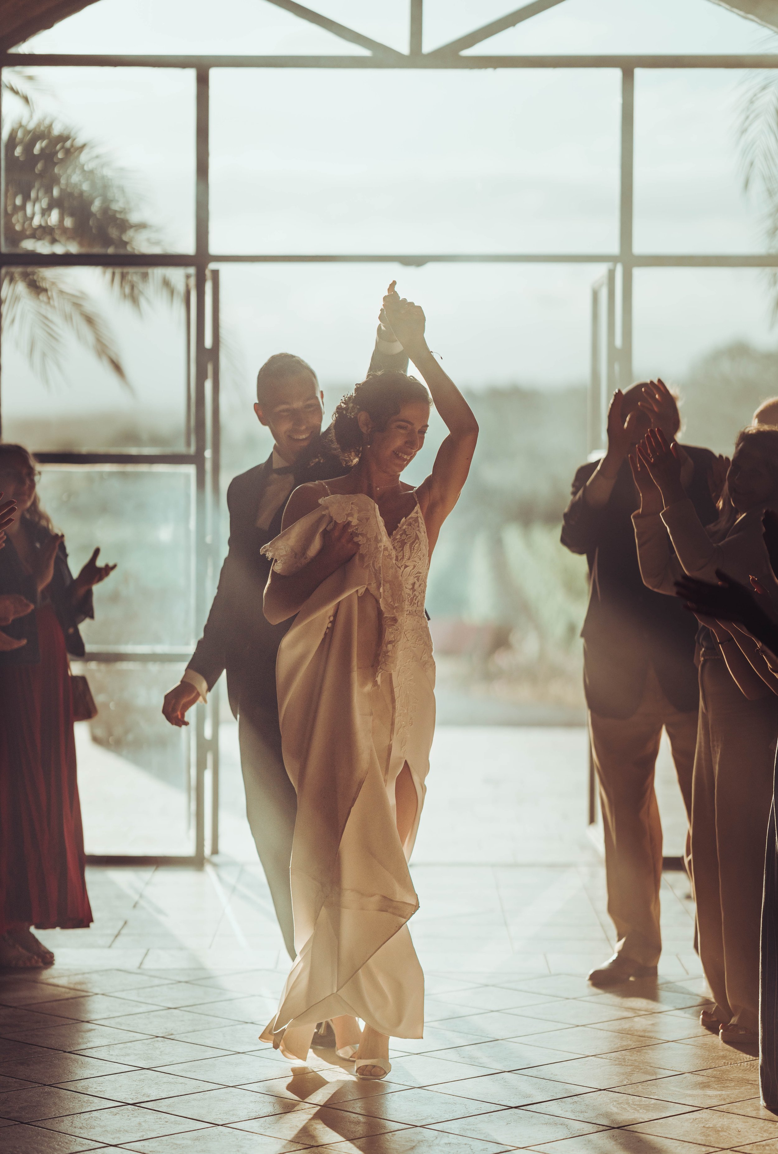 Un couple en mariage danse, souriant, sous une lumière douce à l'entrée d'une salle de mariage proche de Béziers, en Occitanie, entouré d'invités applaudissant.