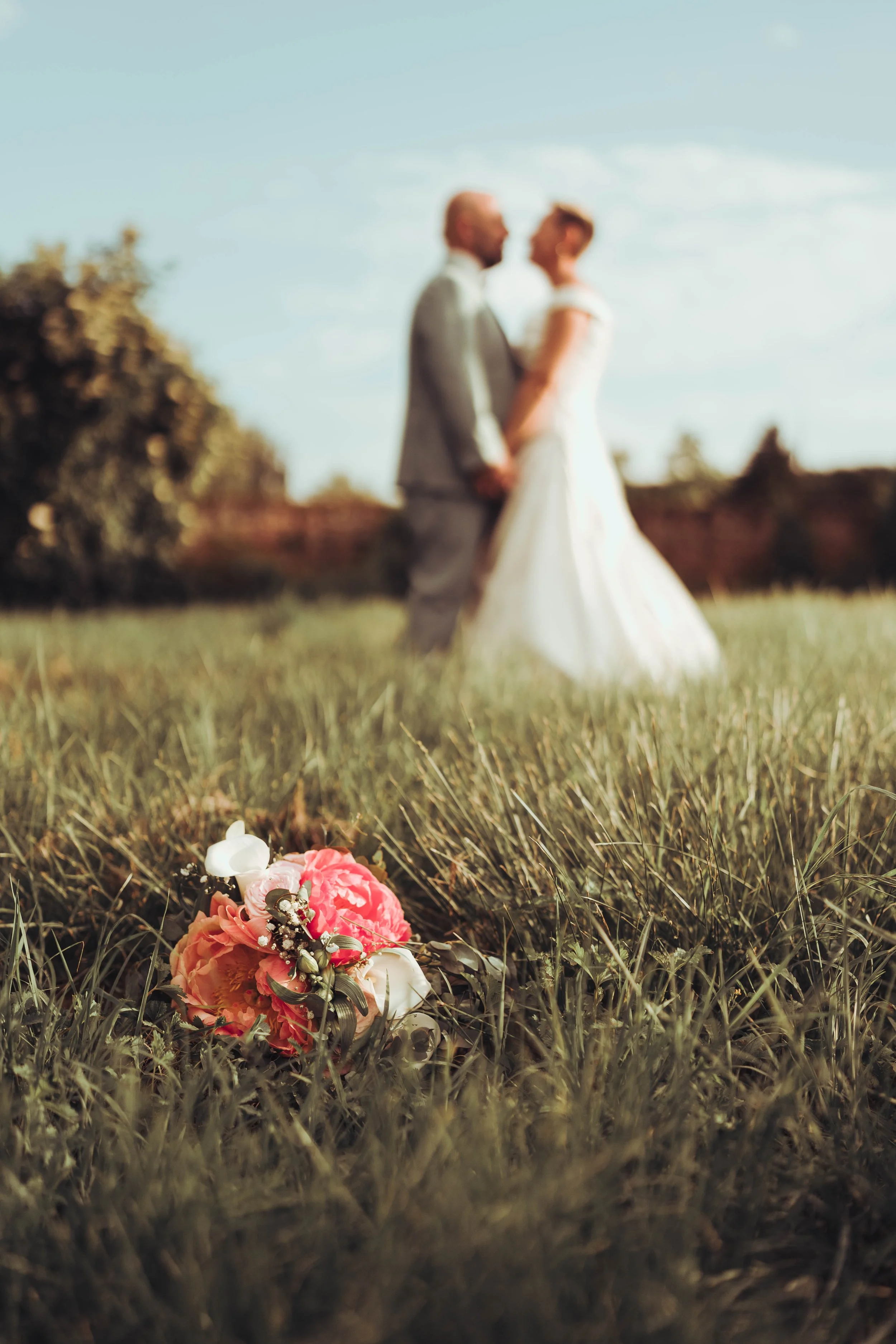 Un bouquet de fleurs rose, blanc et beige posé dans l'herbe devant un couple de mariés flou en arrière-plan dans un domaine de mariage proche de Toulouse.