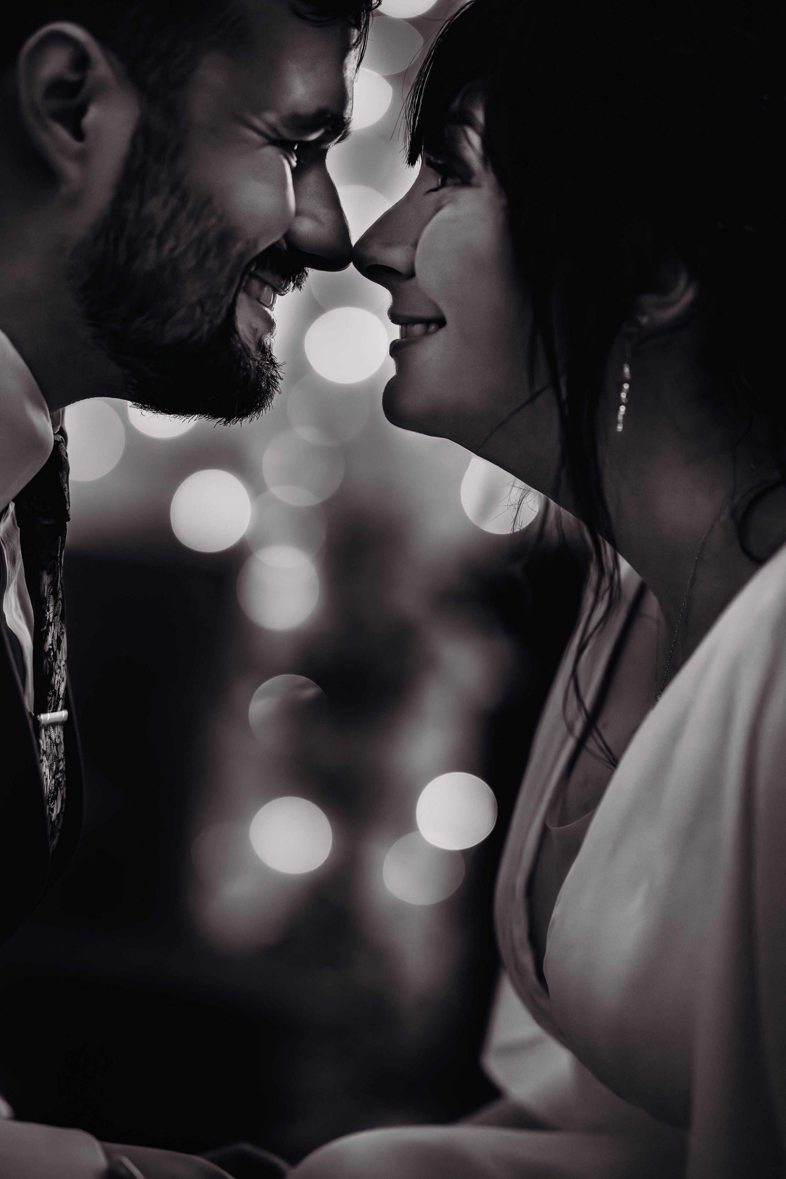Un couple souriant se regarde de près dans une photographie en noir et blanc, avec des lumières floues en arrière-plan au domaine de Gailhaguet, proche de Toulouse.