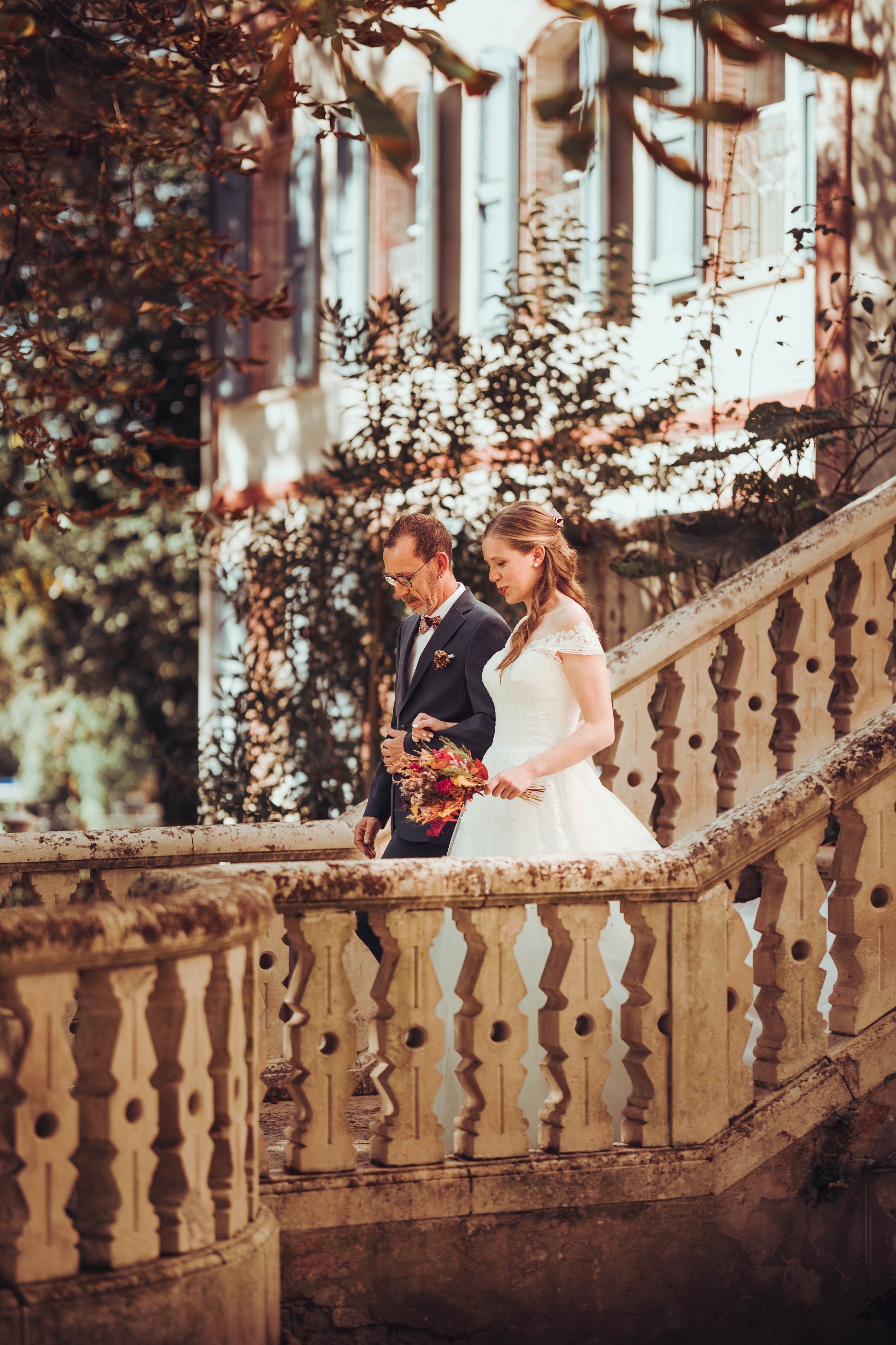 Une mariée en robe blanche et son père en costume noir descendent des escaliers en pierre ornés de balustrades en pierre. Elle se rend à sa cérémonie laïque dans les jardins du manoir du Thouron. 