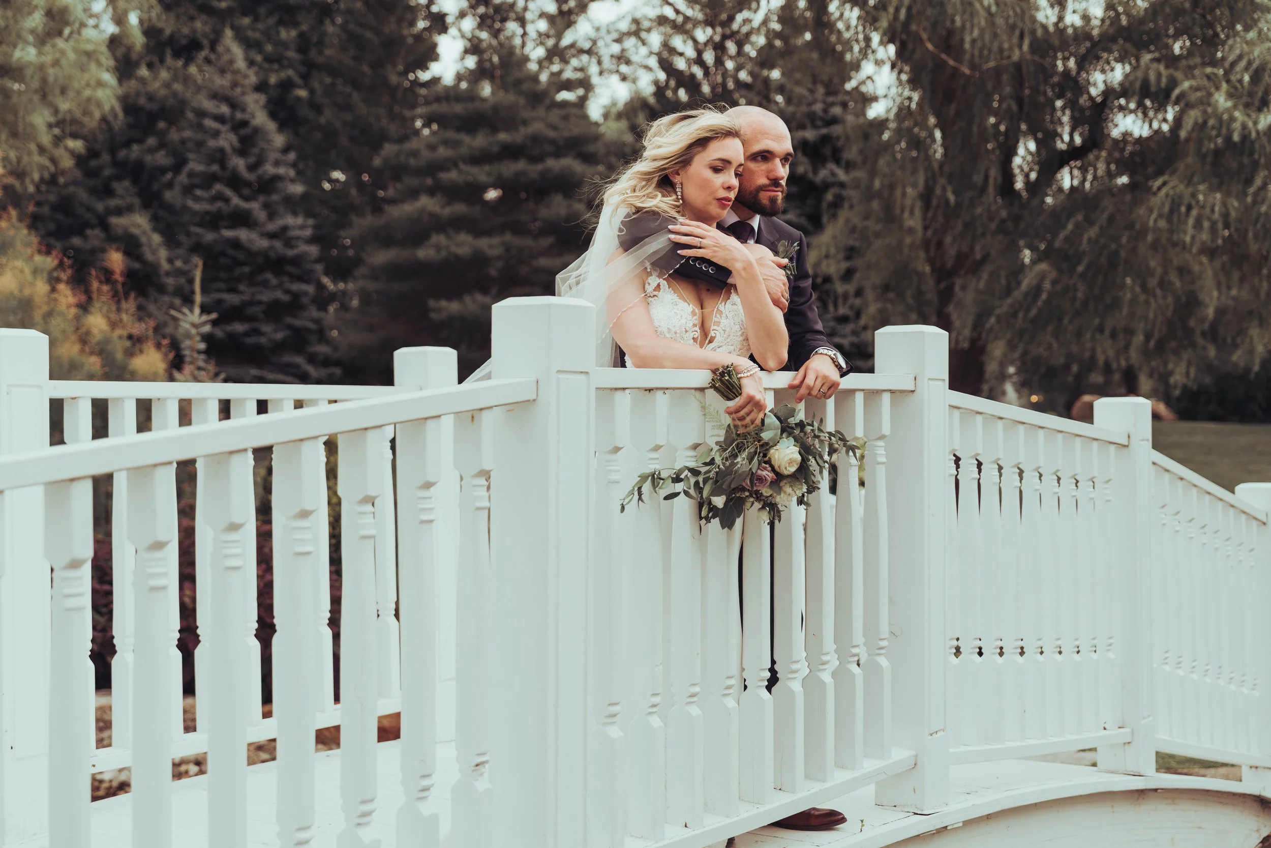 Un couple de mariés au château Vaudreil, proche de Montreal au Canada. La femme en robe de mariage et l'homme en costume, se tient sur un pont blanc en bois dans un parc, avec un bouquet de fleurs à la main, lors d'un mariage en plein air.
